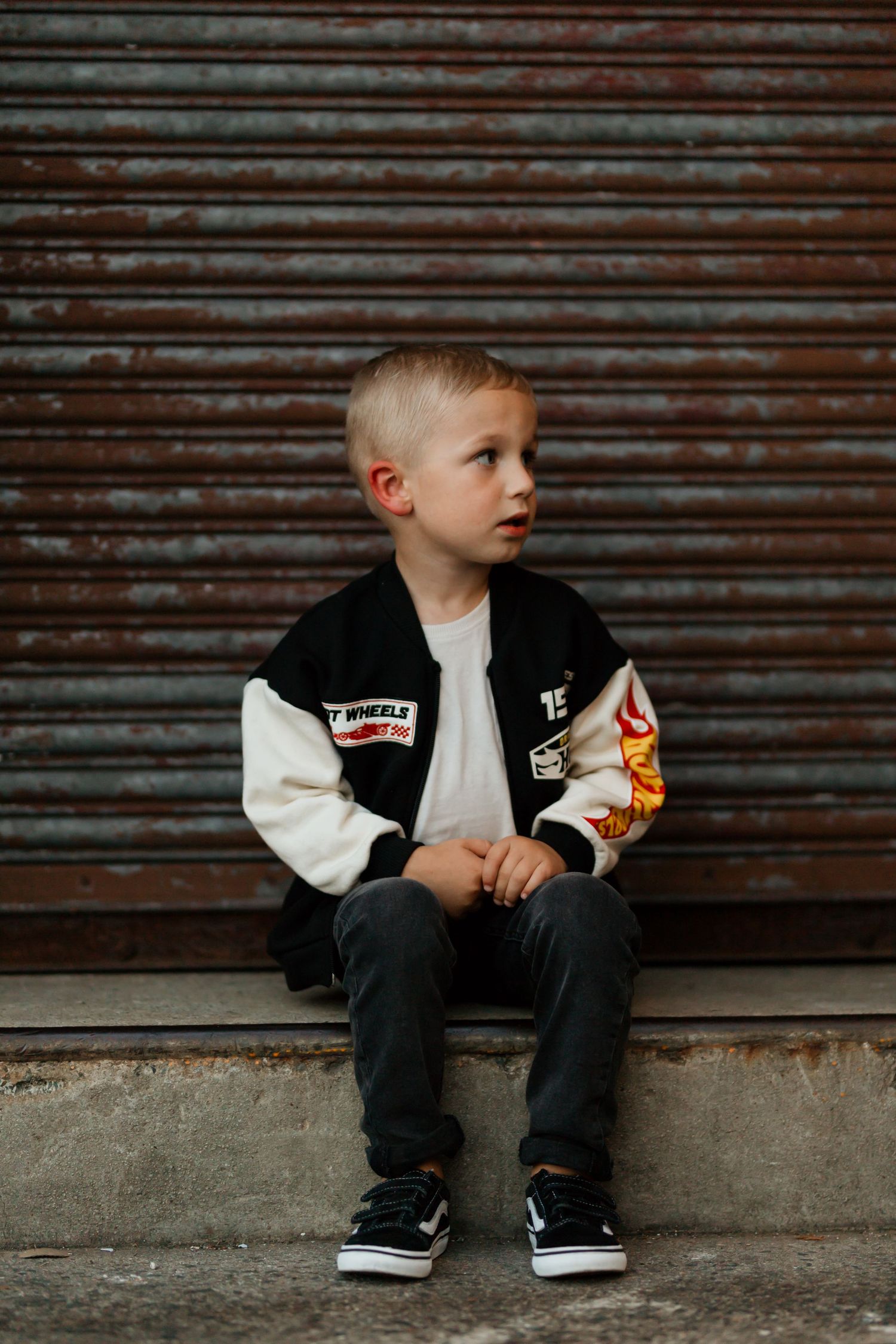 Young boy in varsity jacket with patches sits on steps against rustic metal shutter backdrop.