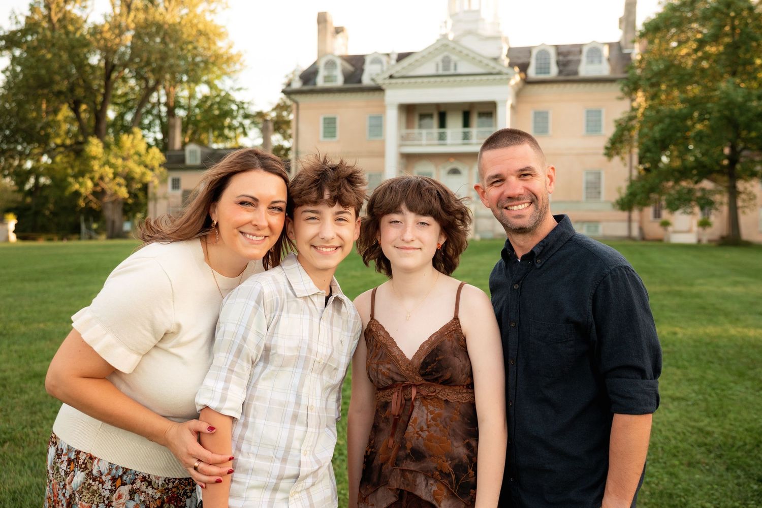 A smiling family of four poses closely together on a sunny green lawn in front of a stately brick mansion surrounded by trees.