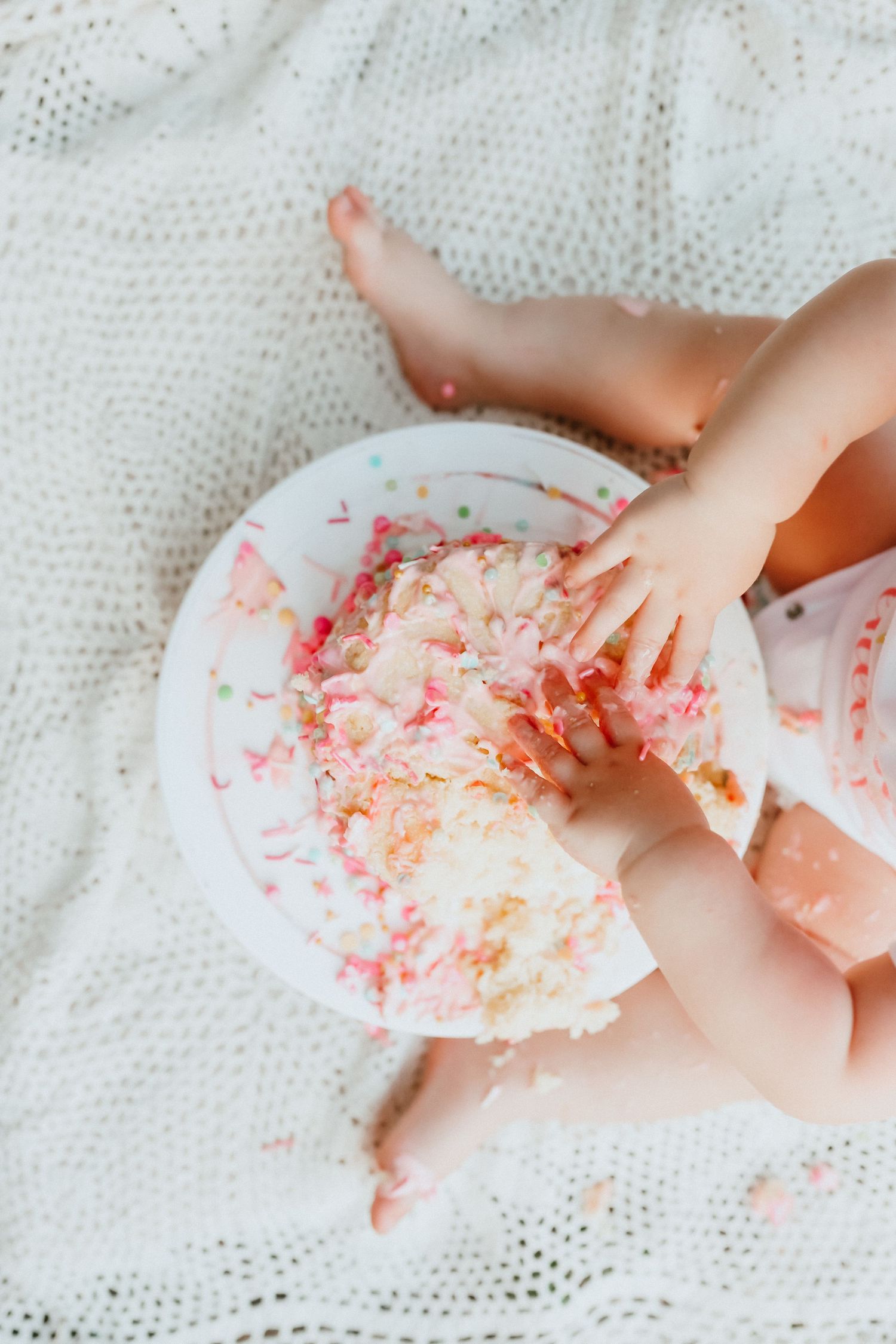 Overhead view of baby hands smashing a pink and white birthday cake on a white blanket.