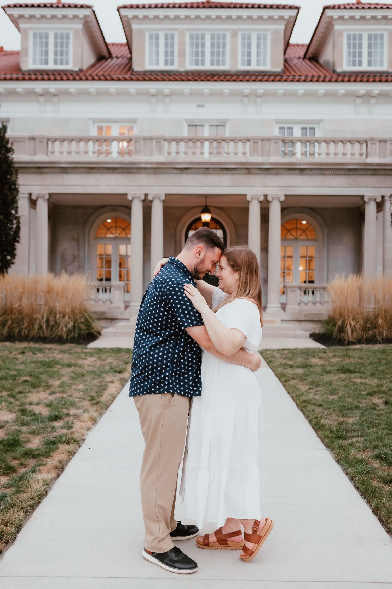 Couple sharing a tender moment on a pathway in front of an elegant mansion at dusk.