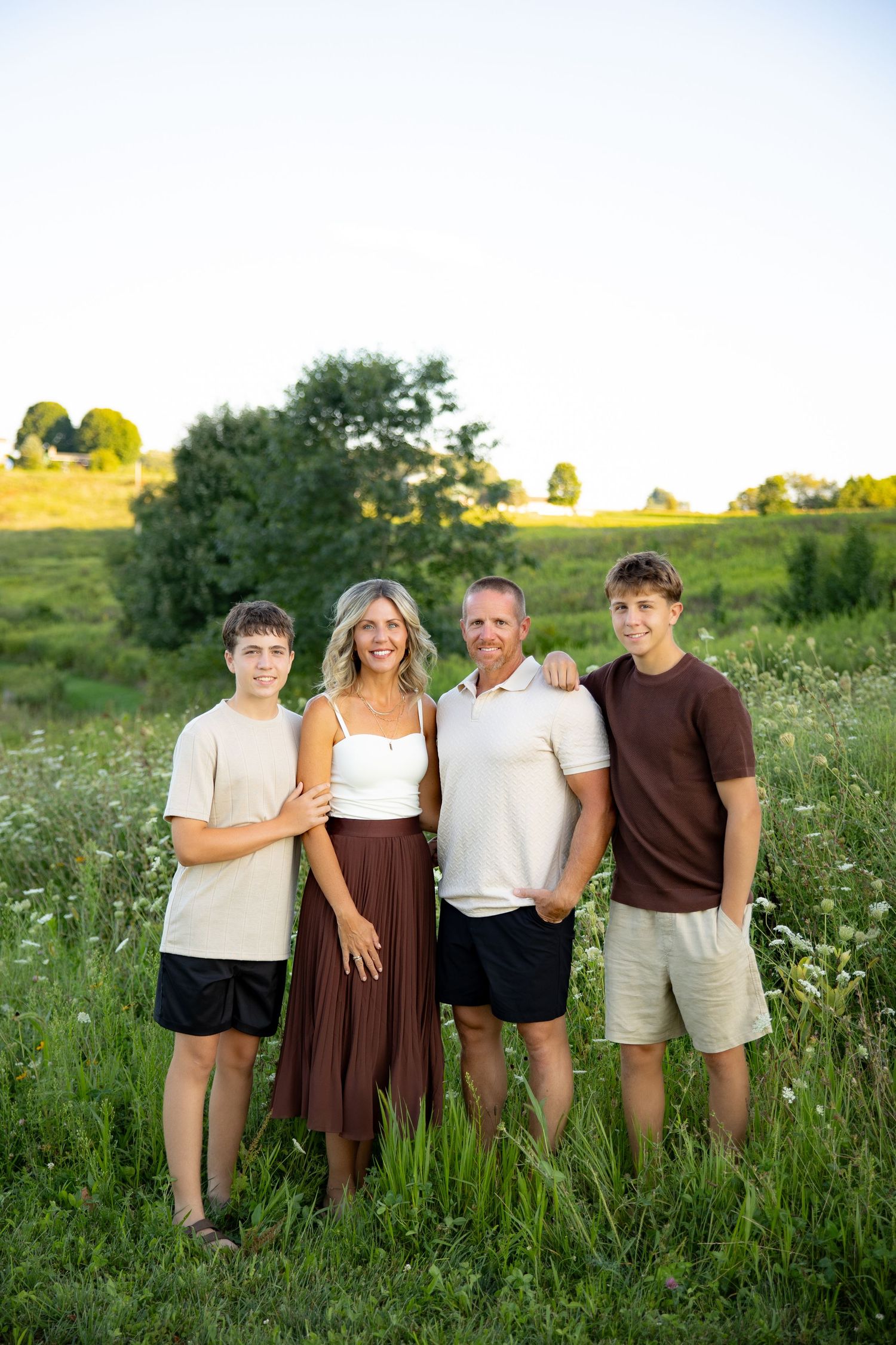Family of four poses in a lush green field at golden hour, wearing coordinated earth-tone outfits.