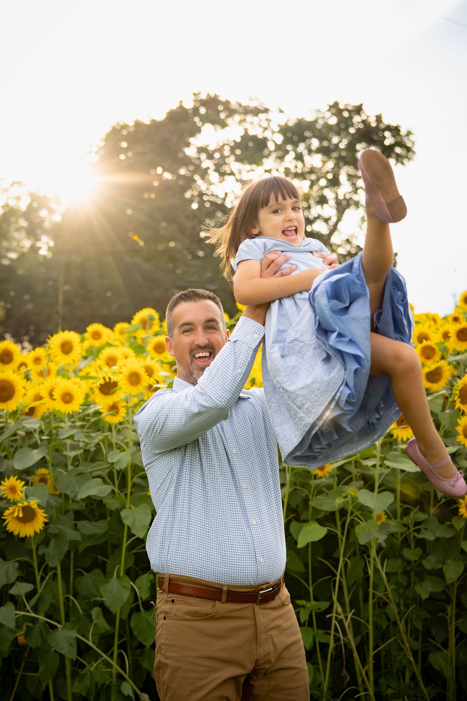 A joyful father lifts his laughing daughter in a sunny sunflower field, both wearing blue outfits.