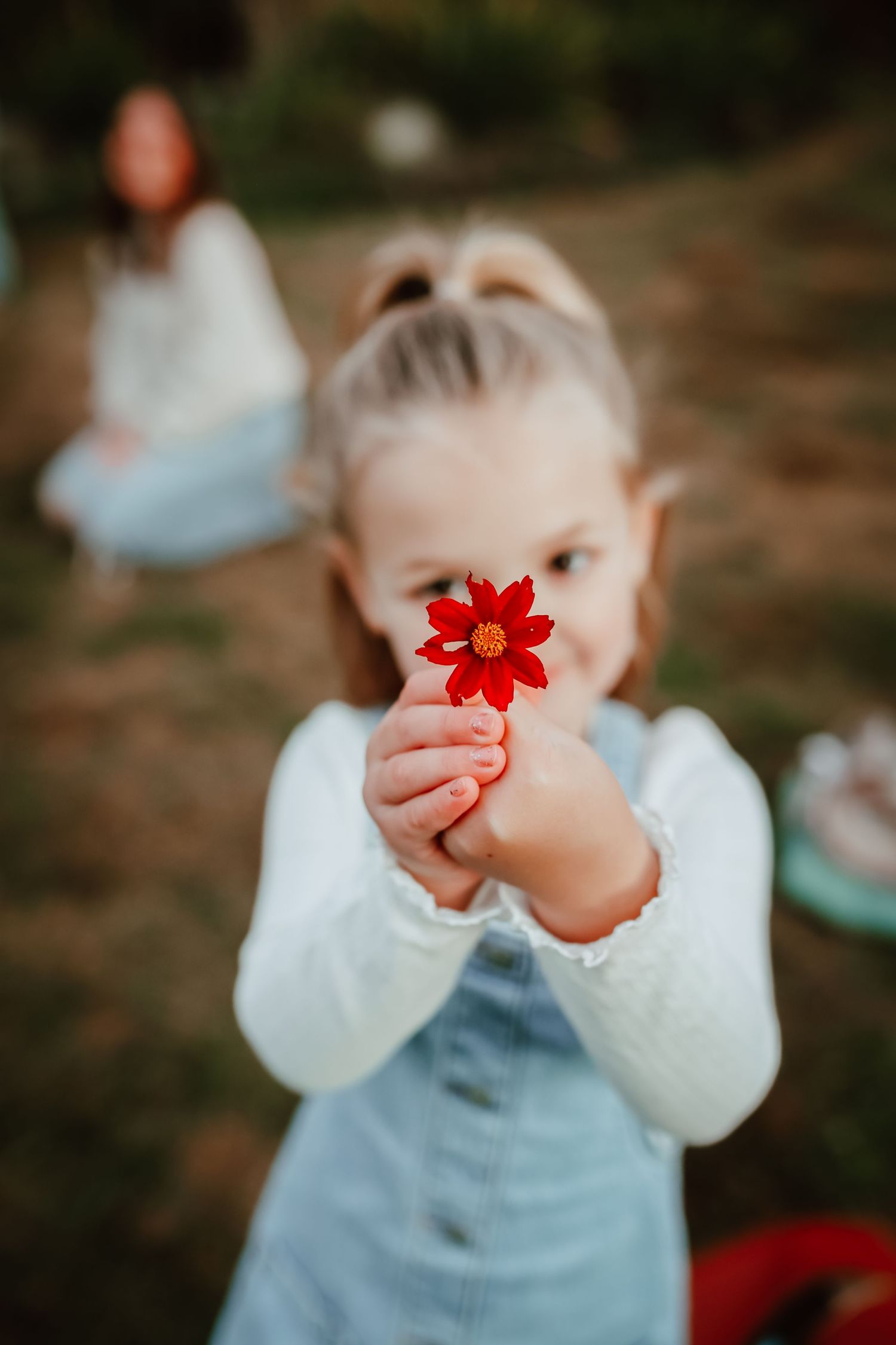A young girl holds a red flower toward the camera in a soft-focus outdoor setting.