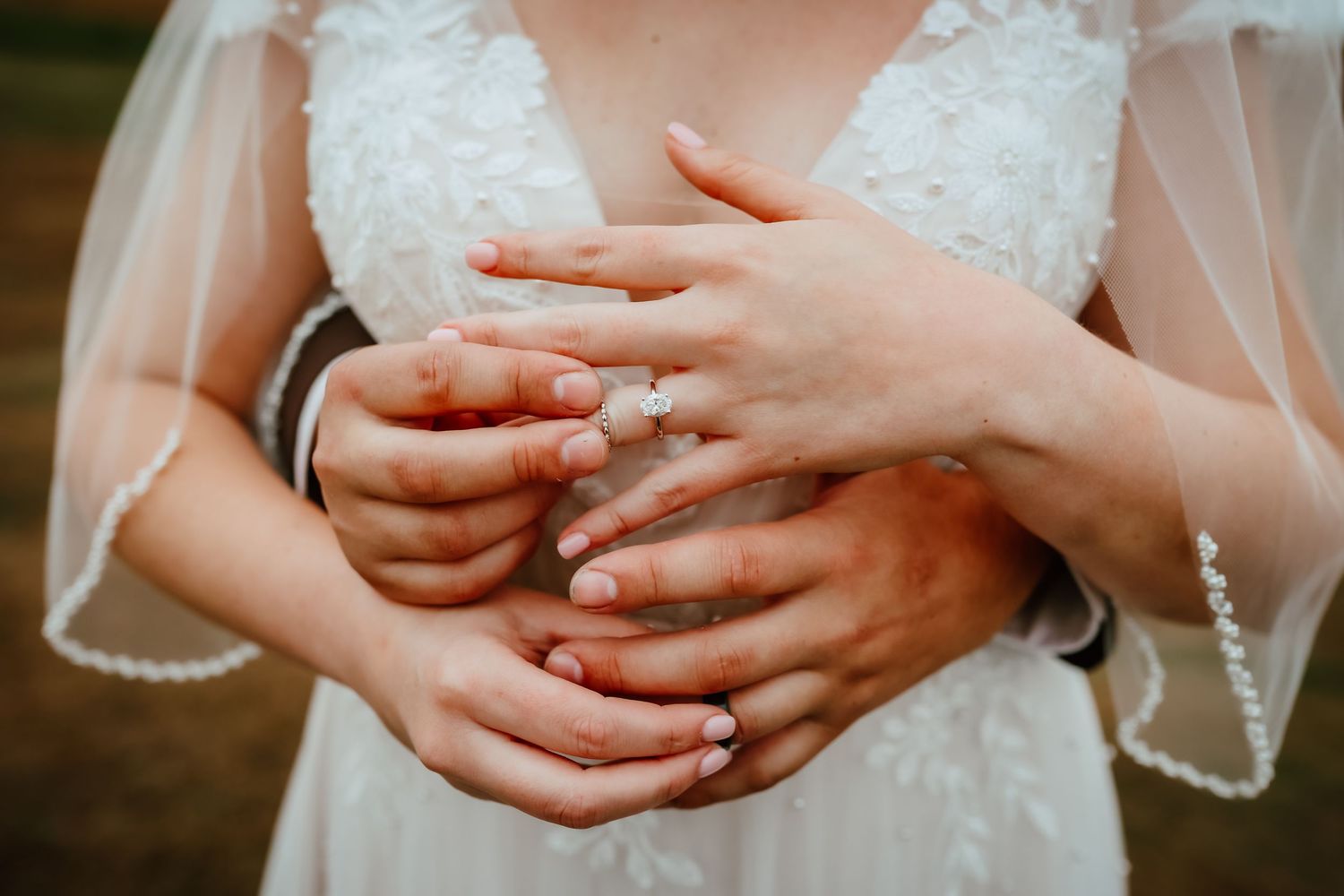 Close-up of bride and groom holding hands together showing wedding rings over bridal gown.