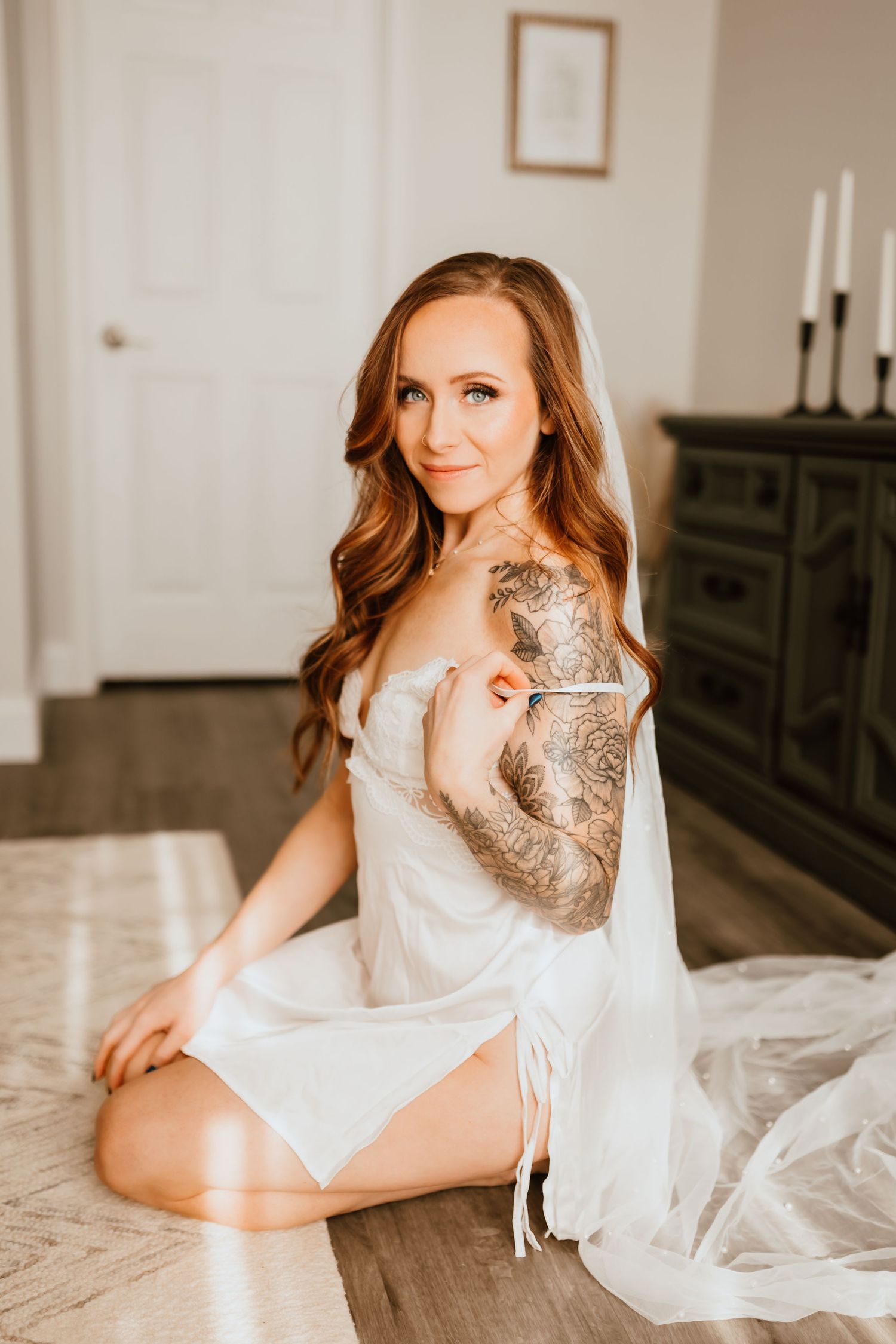 Smiling woman with tattoo sleeve in white bridal lingerie and veil sits on floor.