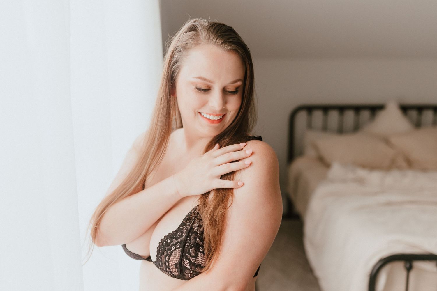 Smiling blonde woman in black lace bra touching her shoulder near sheer curtains in a bedroom.