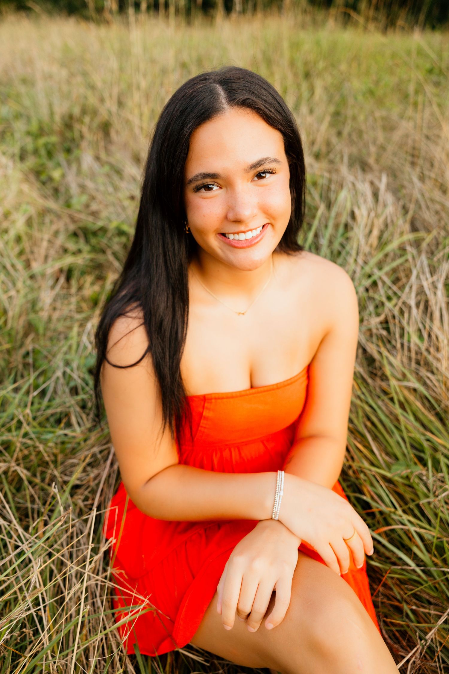 Smiling teen girl in orange strapless dress sits in tall golden grass for outdoor senior portrait.