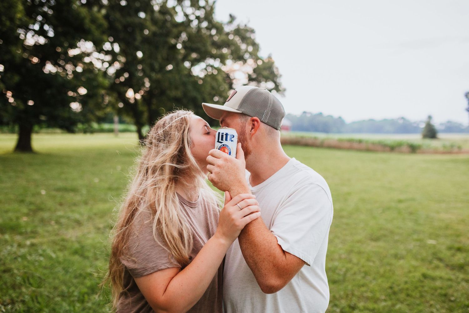 Couple sharing a romantic kiss while holding a beer can in a lush green field outdoors.