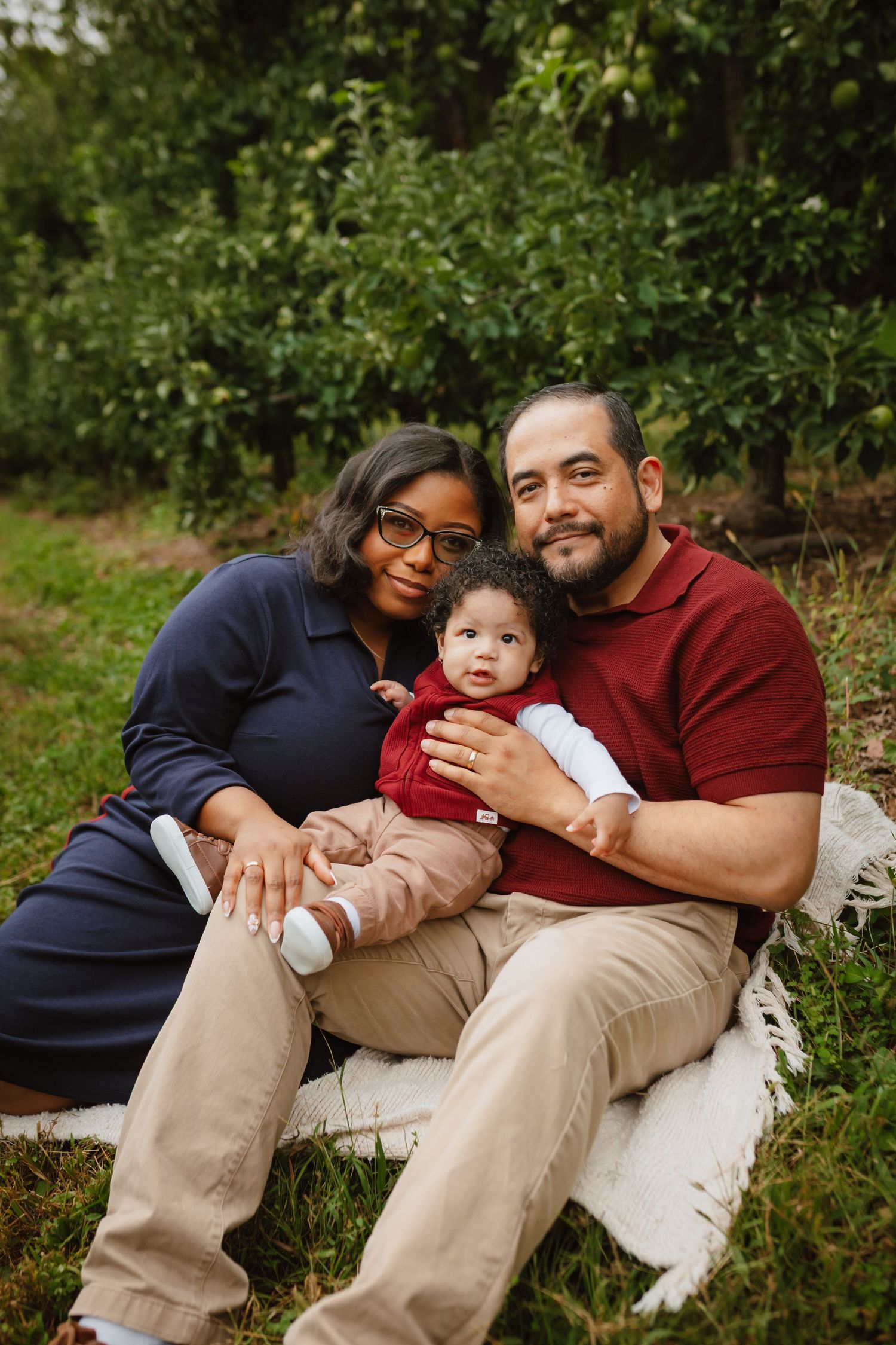 A smiling couple sits on a cream blanket in an orchard, holding their baby between them in a warm outdoor family portrait.