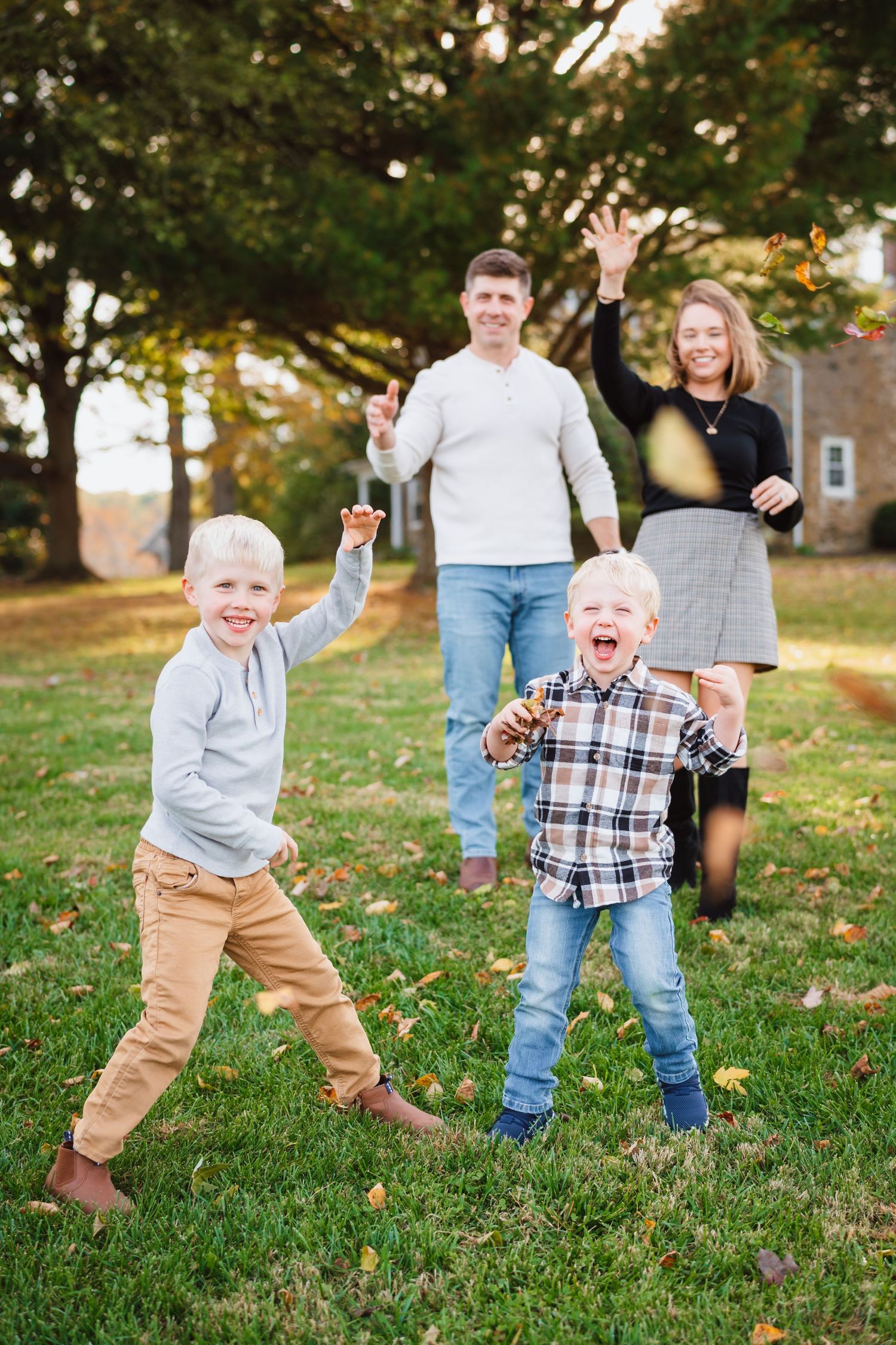 Joyful family of four playing in autumn park, two excited boys in foreground with parents waving behind them.