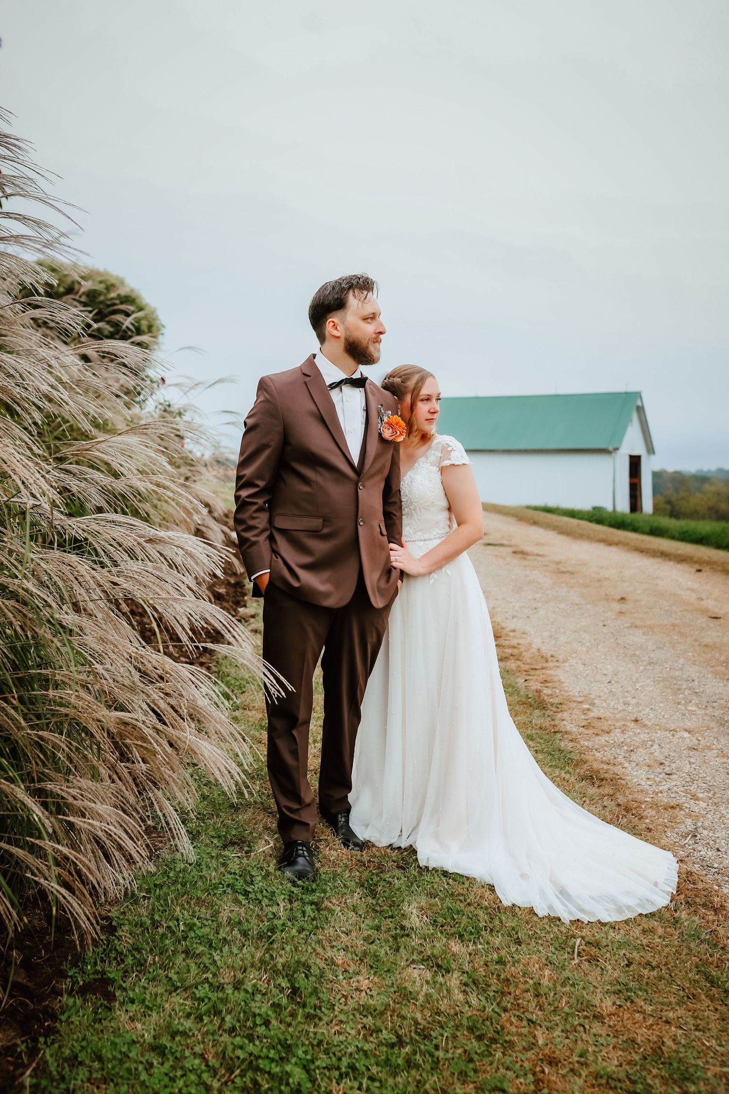 Bride and groom posing together on rural dirt path beside tall pampas grass.