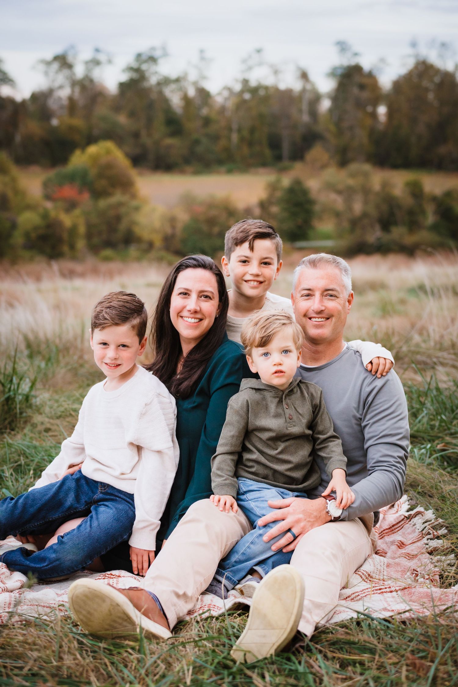 Family of five sitting on blanket in autumn field, parents with three young boys smiling during outdoor fall photo session.