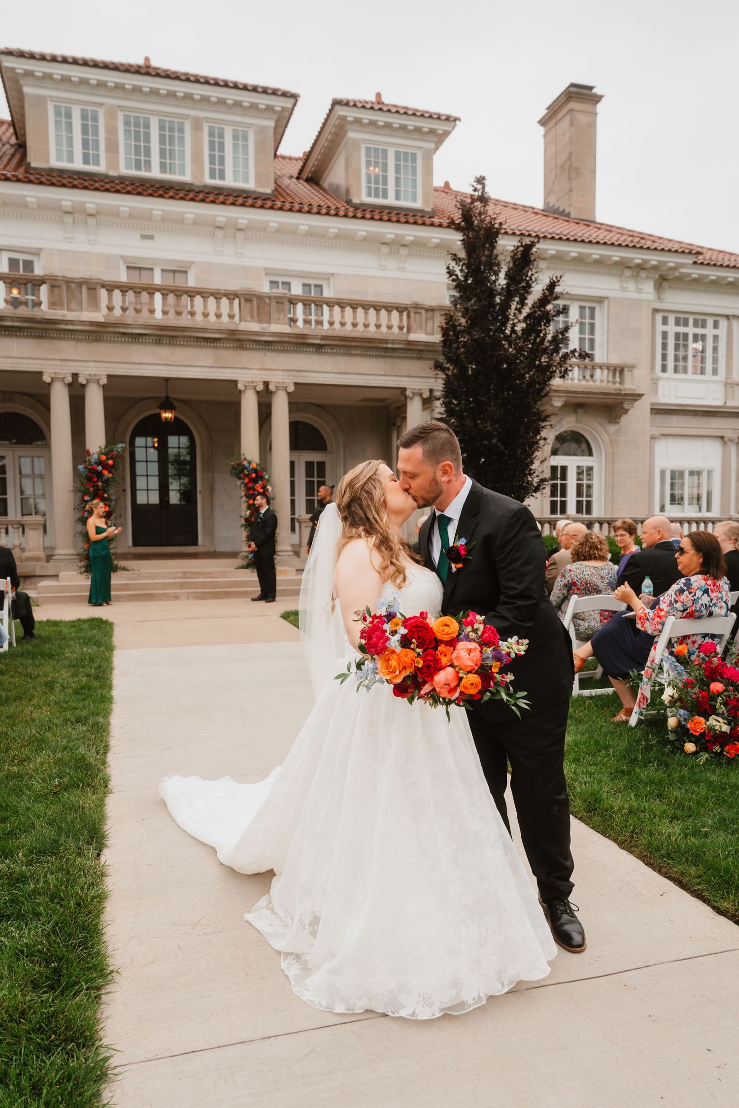 Bride and groom share first kiss as newlyweds outside grand estate with vibrant bridal bouquet.