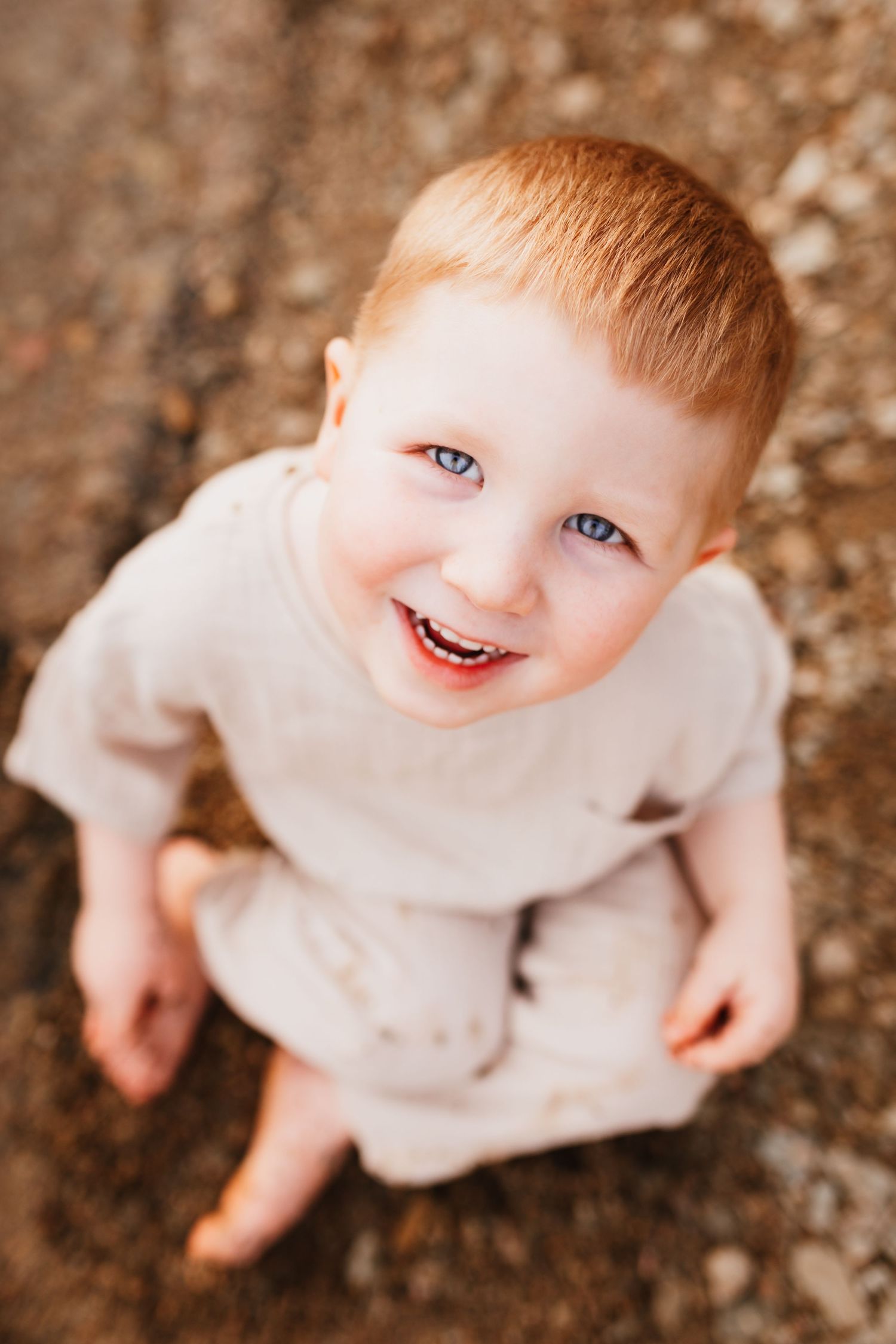 Smiling red-haired toddler boy with blue eyes looking up at camera outdoors.