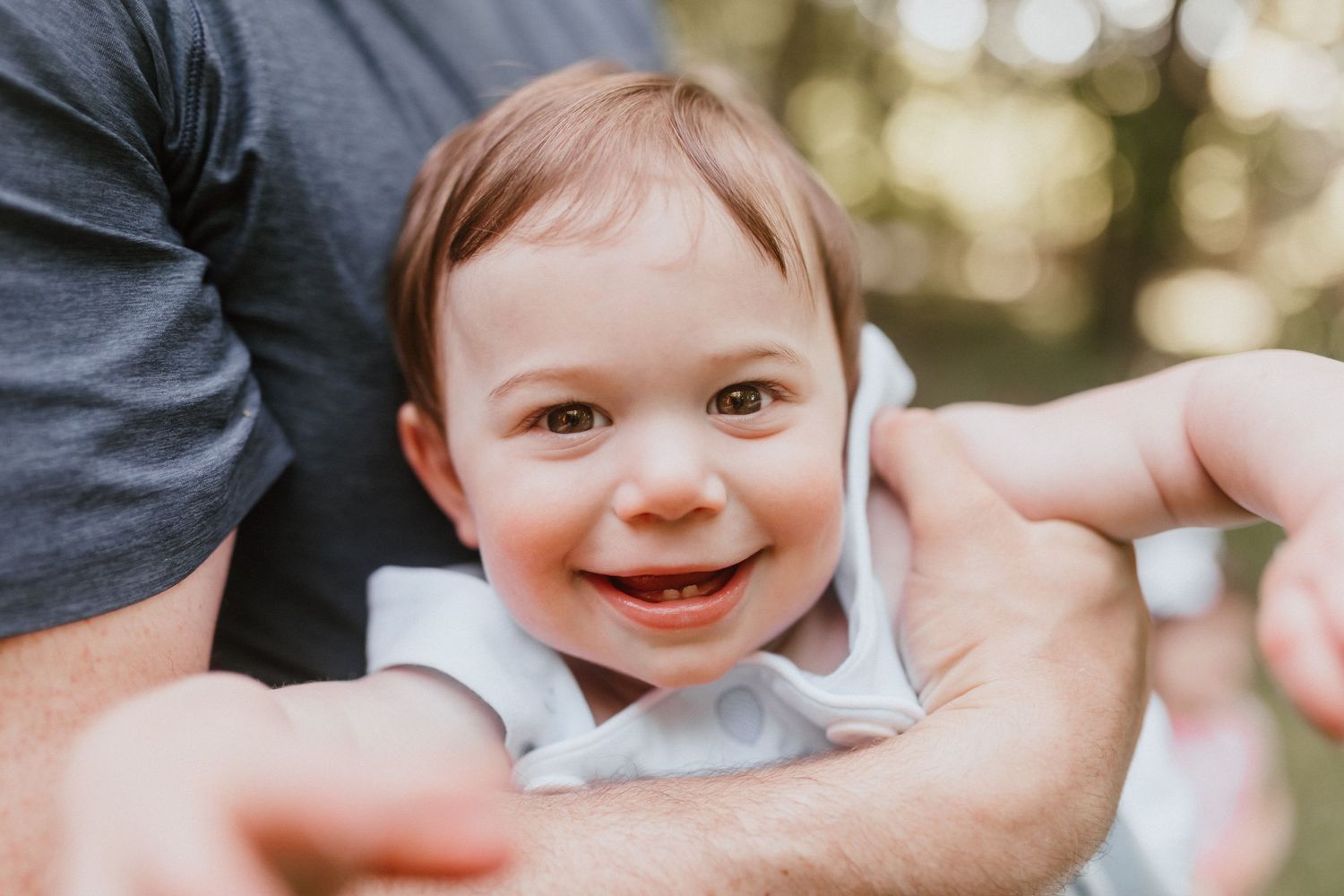Smiling baby with two teeth being held by adult hands in sunlit outdoor setting.