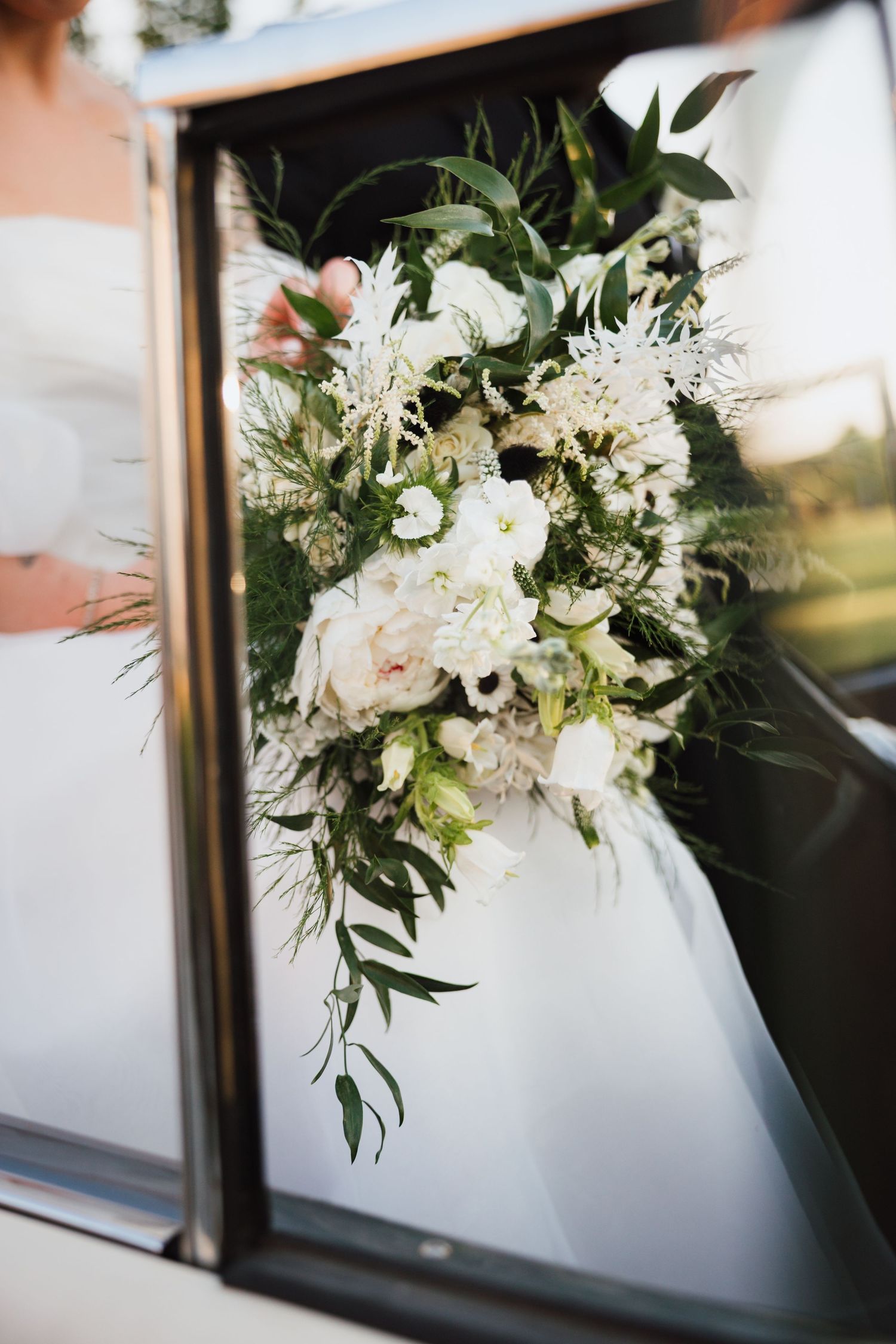 Lush white and blush bridal bouquet with greenery reflected in vintage car window at sunset.