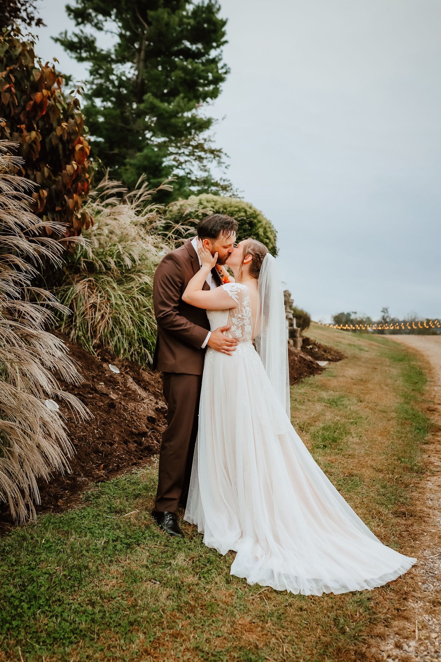 Bride and groom kissing beside tall ornamental grasses on overcast wedding day.