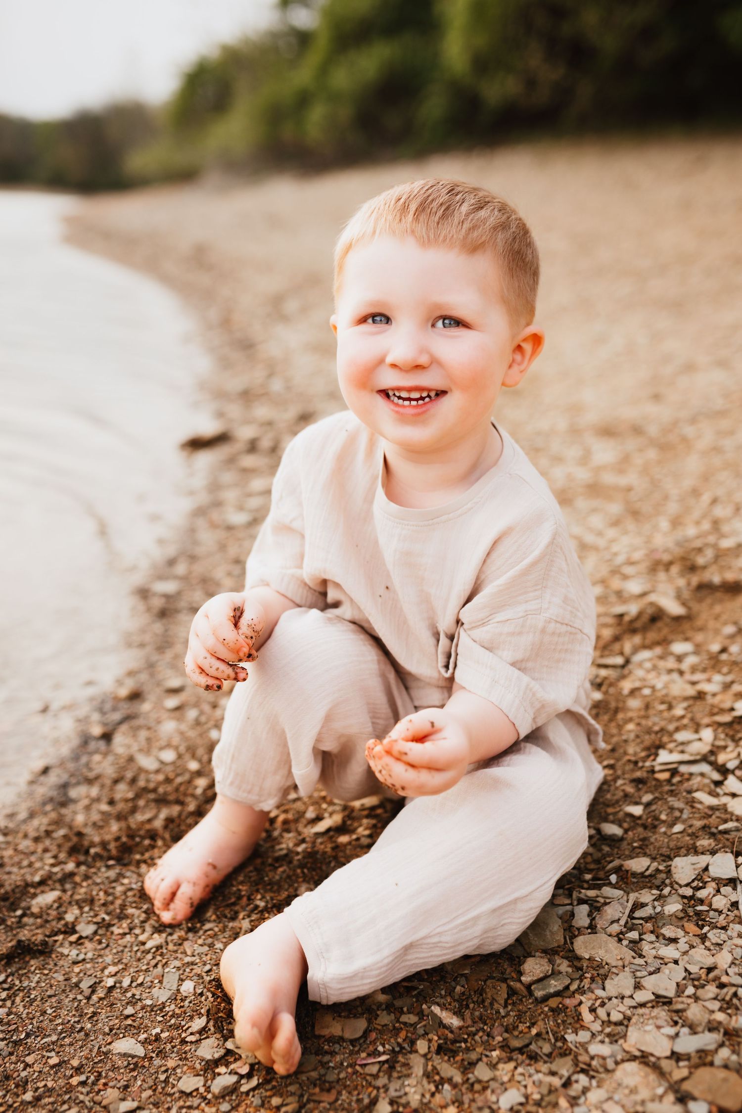 Smiling toddler in linen outfit sitting barefoot on a sandy beach shoreline.