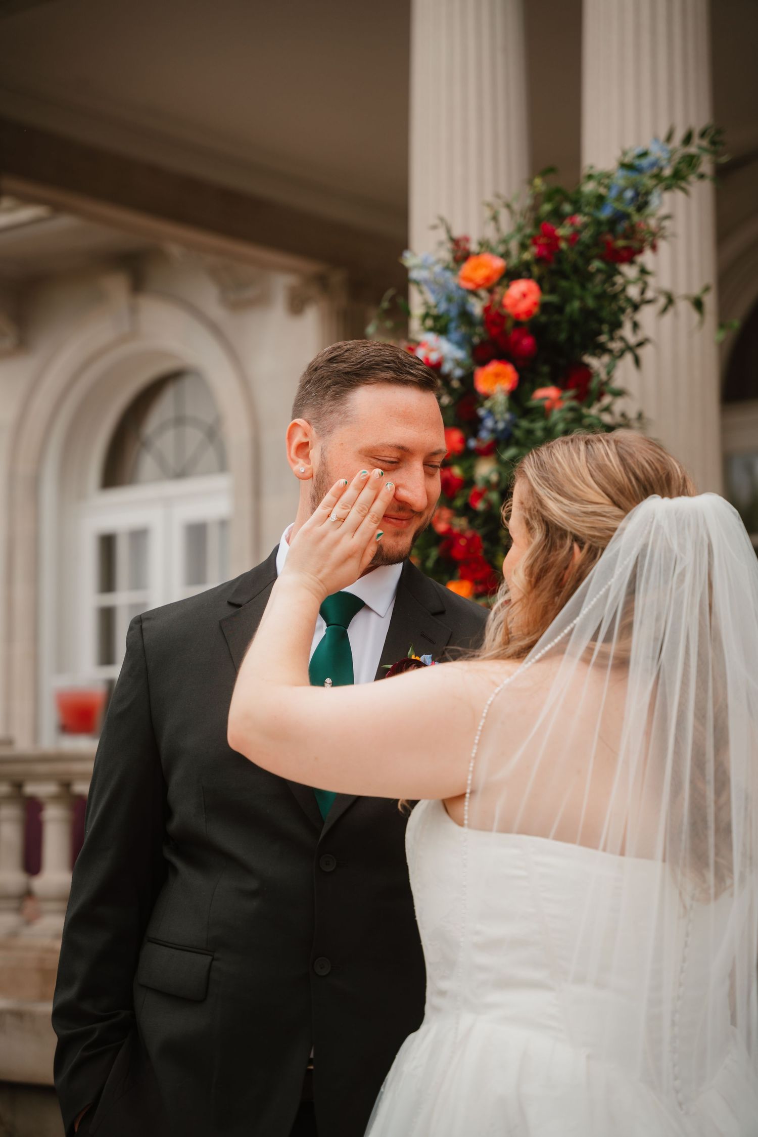 Bride gently wipes tear from emotional groom's face during first look outside elegant venue.