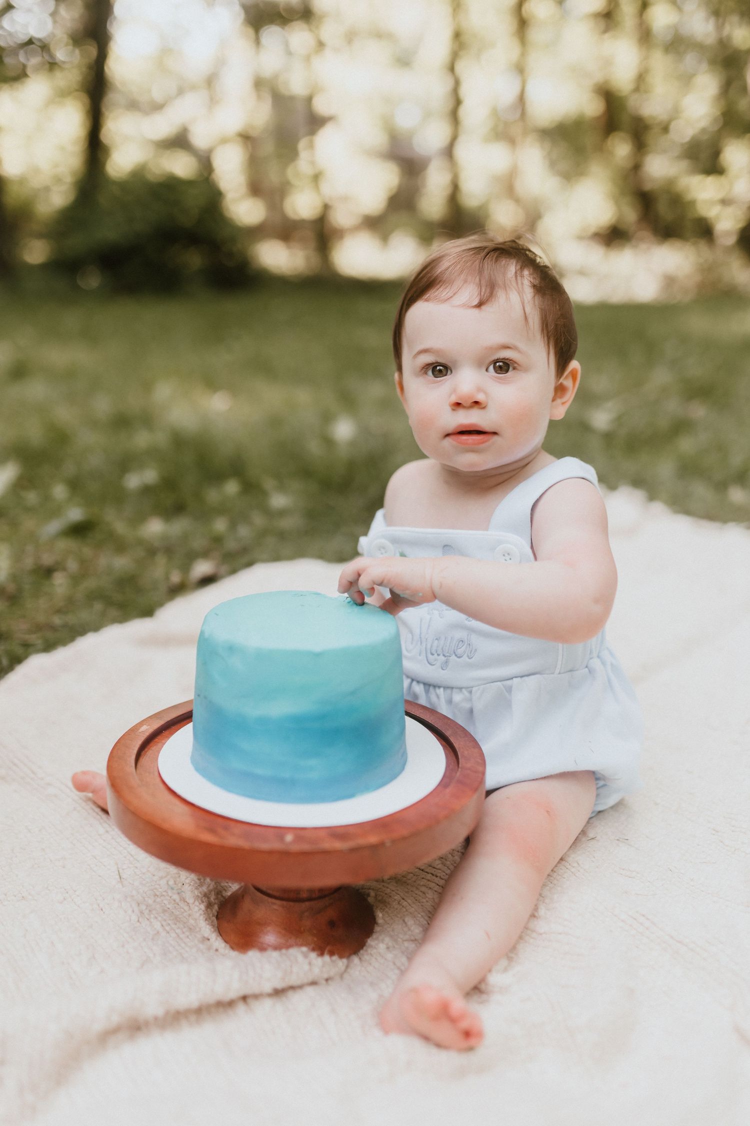 Baby in white romper touches blue ombre first birthday smash cake on wooden stand outdoors.