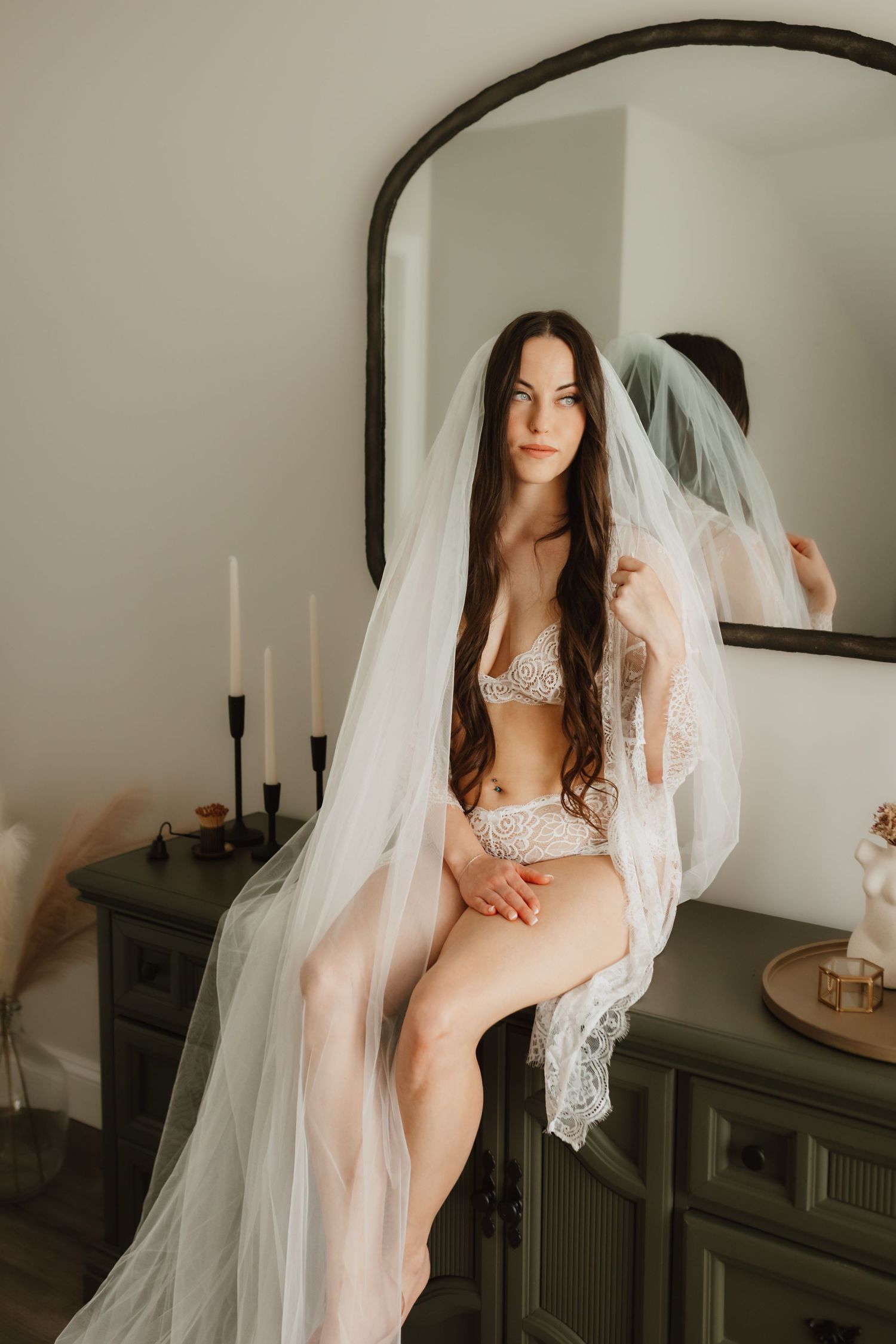 Brunette woman in white lace lingerie sitting on green dresser draped in tulle veil near mirror.