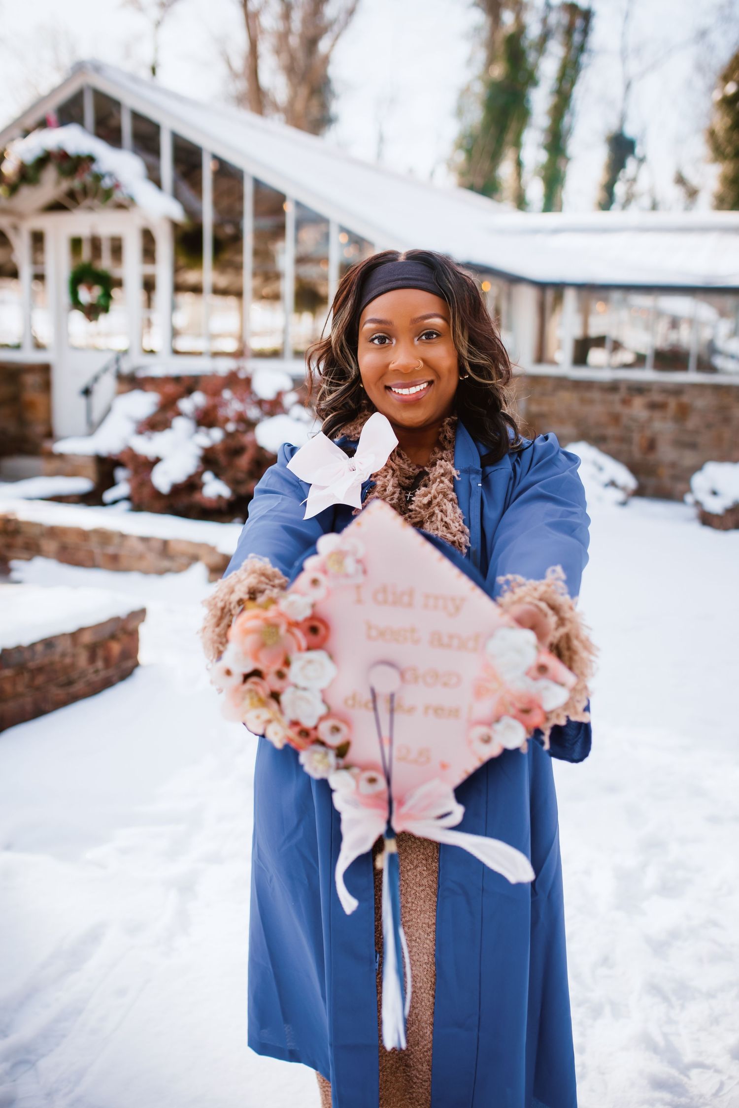 Graduate in blue gown holds decorated pink mortarboard cap in snowy outdoor setting.