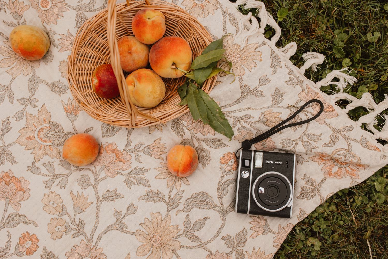 Flat lay of peaches in wicker basket with vintage instant camera on floral blanket outdoors.