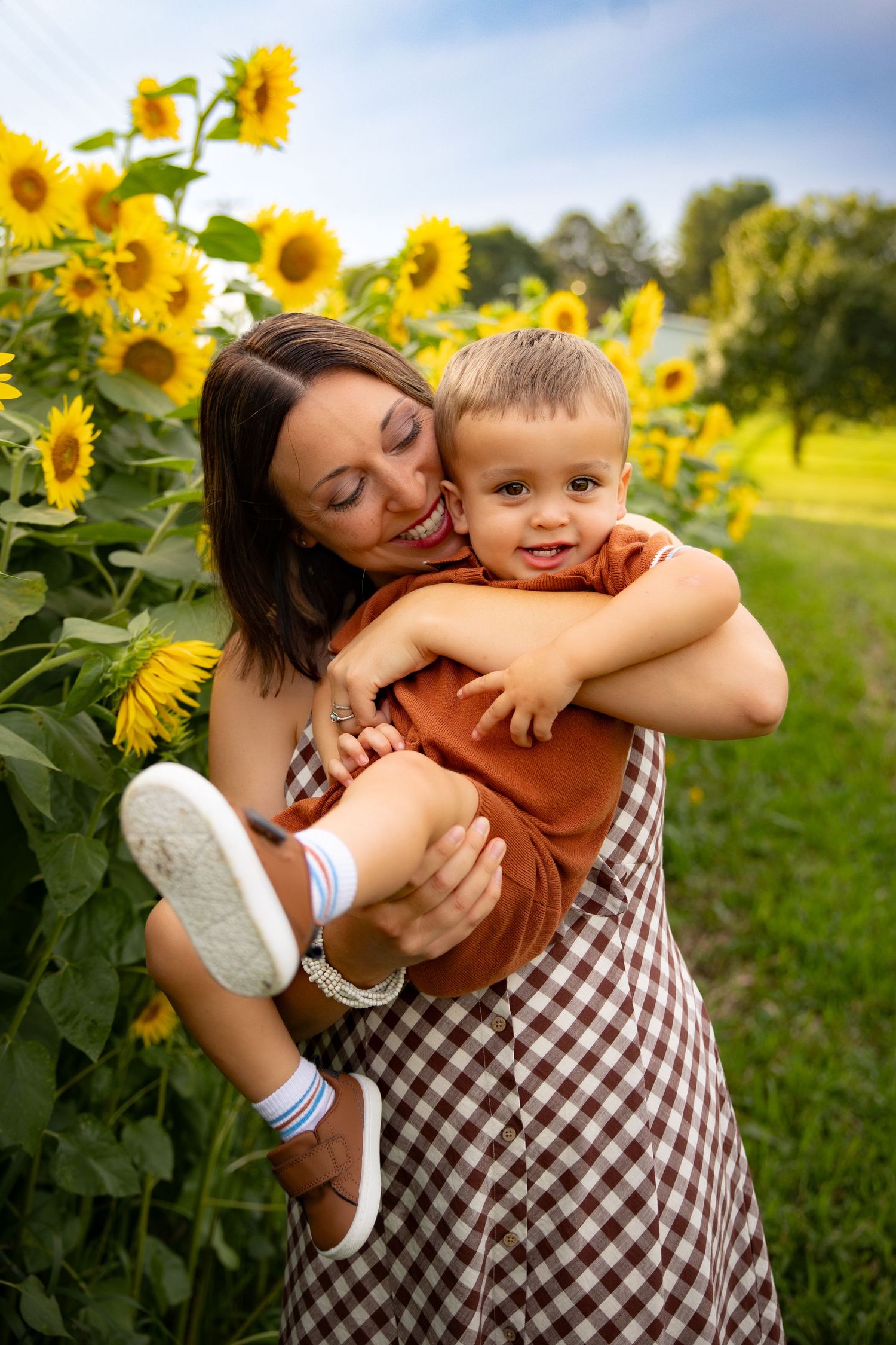 A smiling mother holds her happy toddler son in a sunflower field, both wearing warm tones on a sunny day.