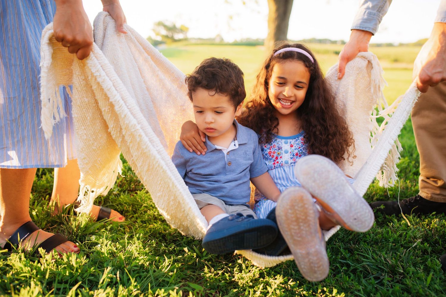 Two children sitting together in a blanket swing held by parents in a sunny green backyard.