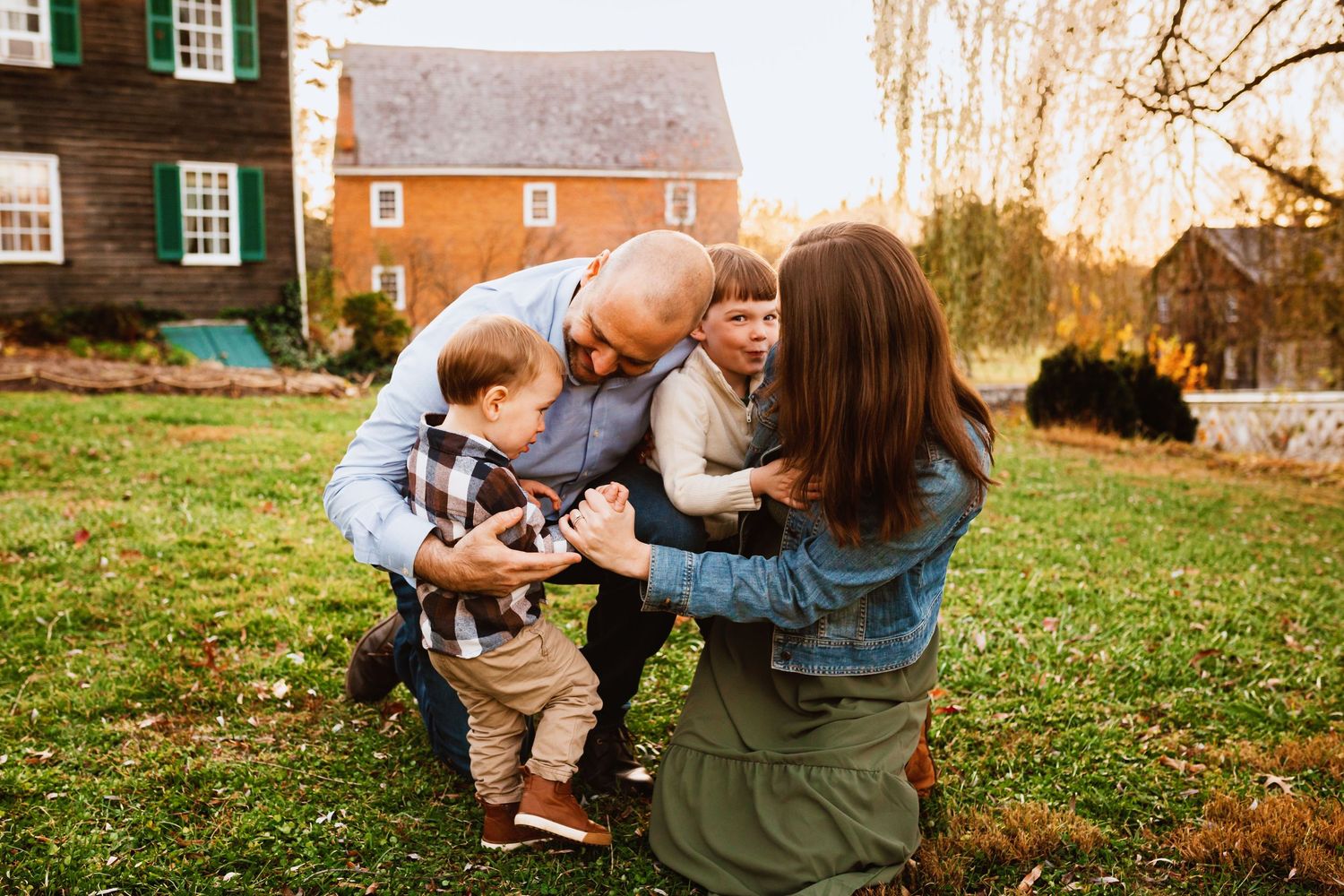 Loving family of four playfully hugging and laughing together on a green lawn near a brick home.