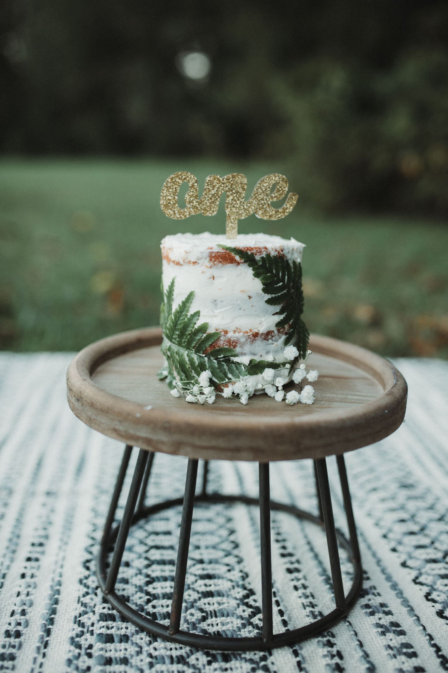 Rustic naked one-tier birthday cake with fern leaves and gold glitter one topper on wooden cake stand.