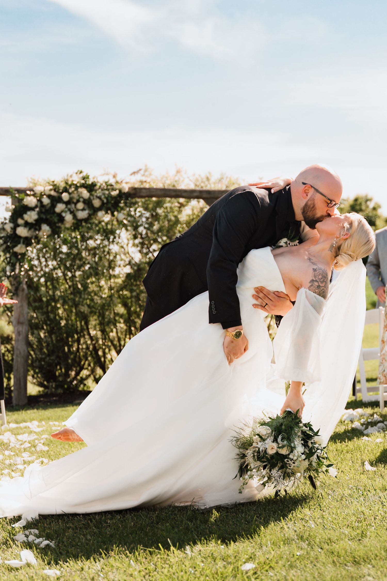Bride and groom sharing romantic dip kiss outdoors at floral wedding arch on sunny day.