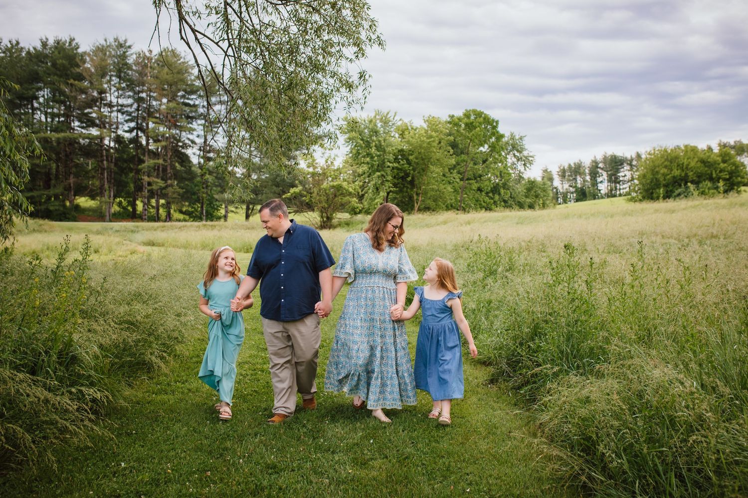 A happy family of four walks through a lush green meadow on a cloudy day.