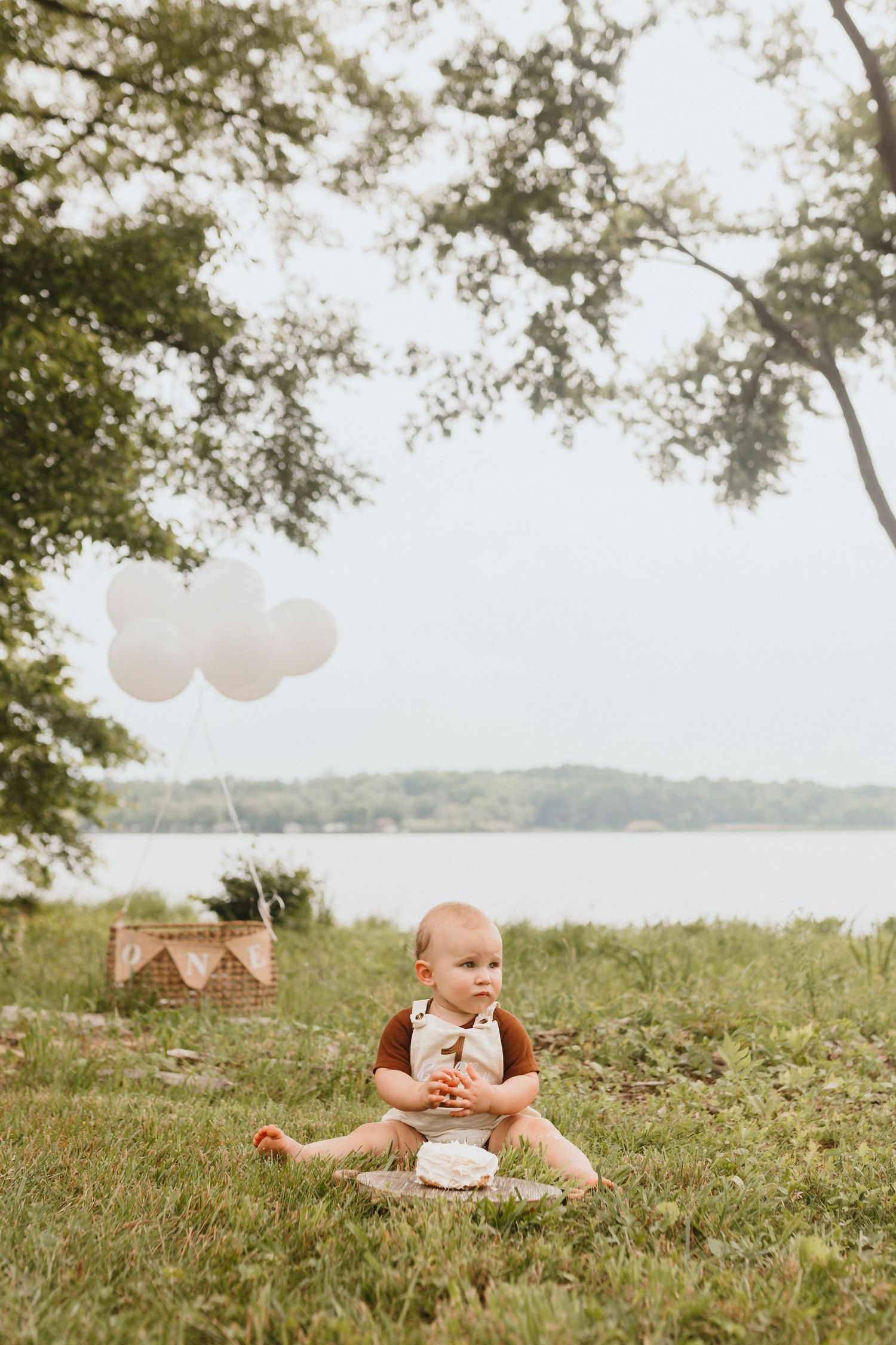 Baby in brown romper sits on grass near lake with white balloons and first birthday cake.