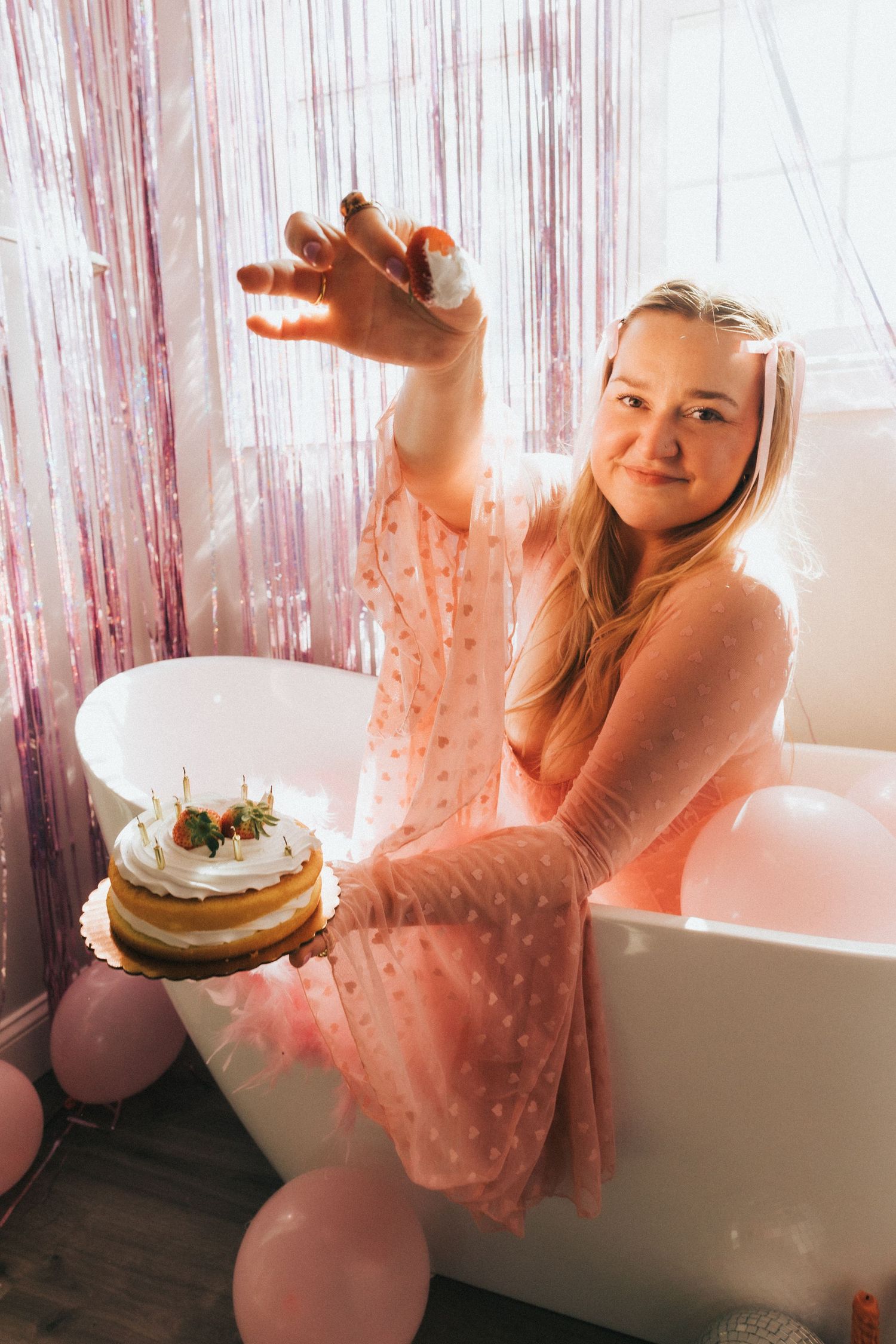 Smiling young woman holds birthday cake while sitting on bathtub edge with pink balloon decorations.
