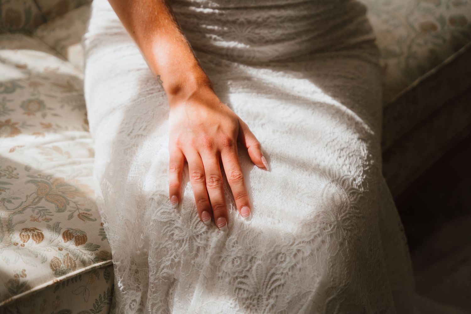 Close-up of bride's hand resting on white lace wedding dress in soft sunlight.