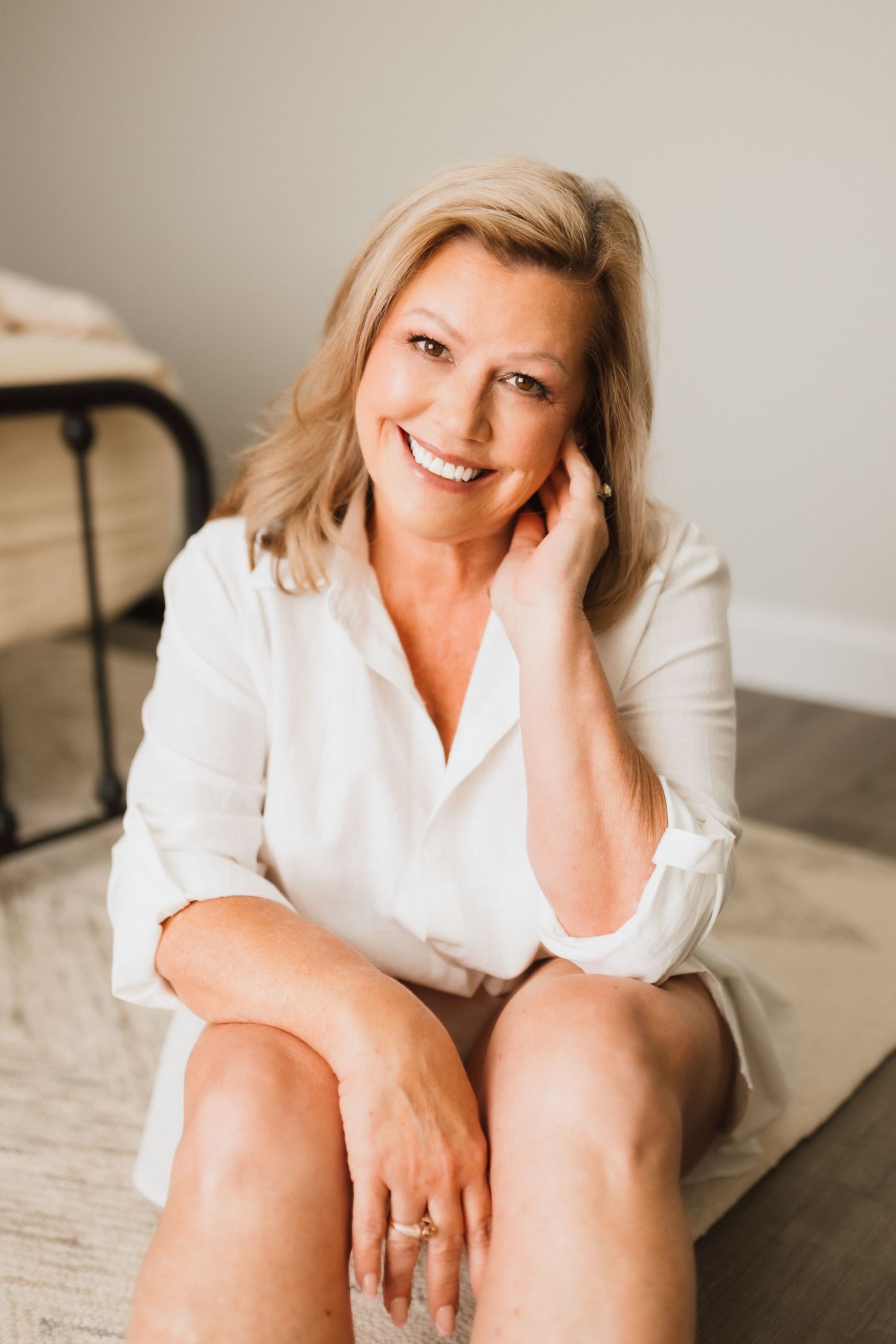 Smiling mature blonde woman in white button-down shirt sits casually on floor.