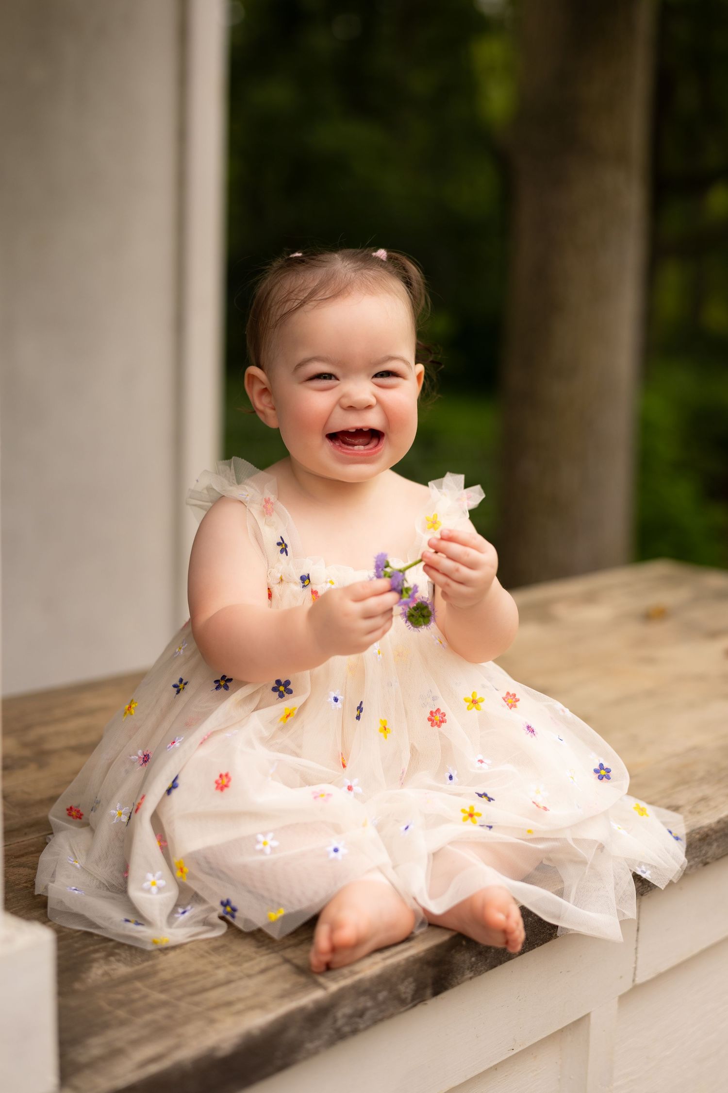 Laughing baby girl in a floral embroidered white dress sitting on a wooden porch railing.