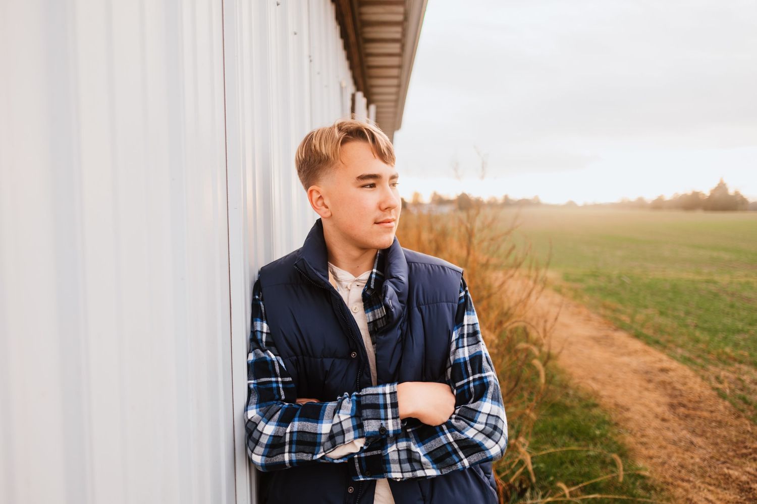 Young man in plaid shirt and vest leaning against white barn wall in an autumn field.