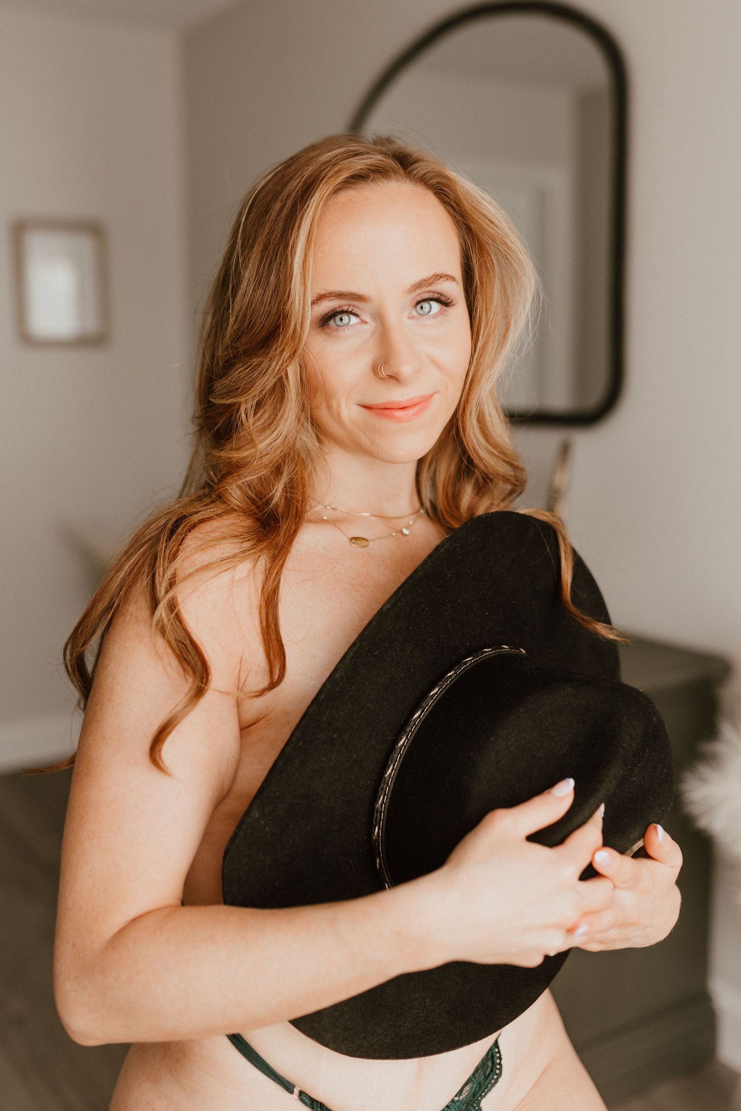 Smiling redhead woman holding a black cowboy hat in a softly lit bedroom setting.