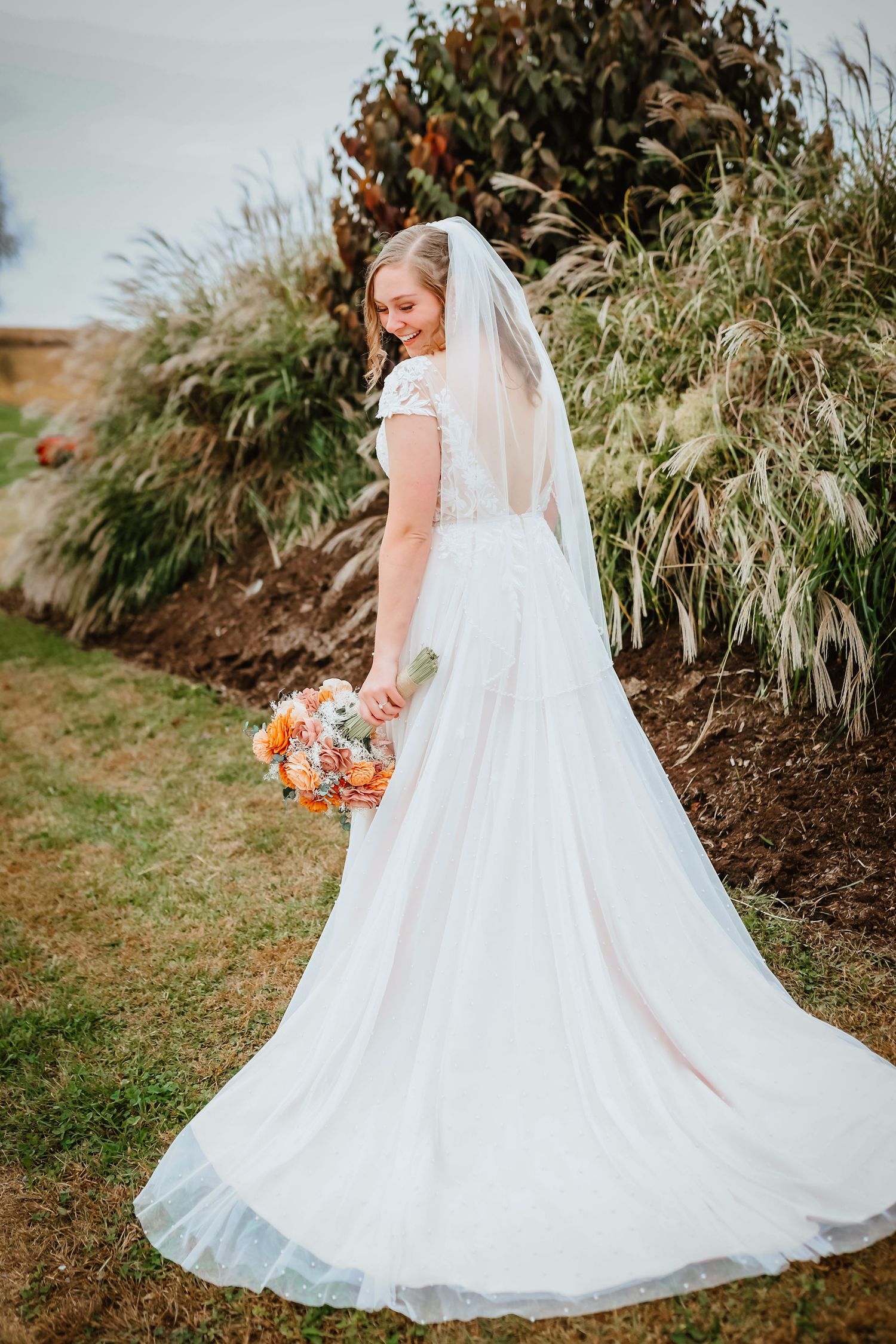 Bride in white lace gown with veil holding orange bouquet viewed from behind outdoors.