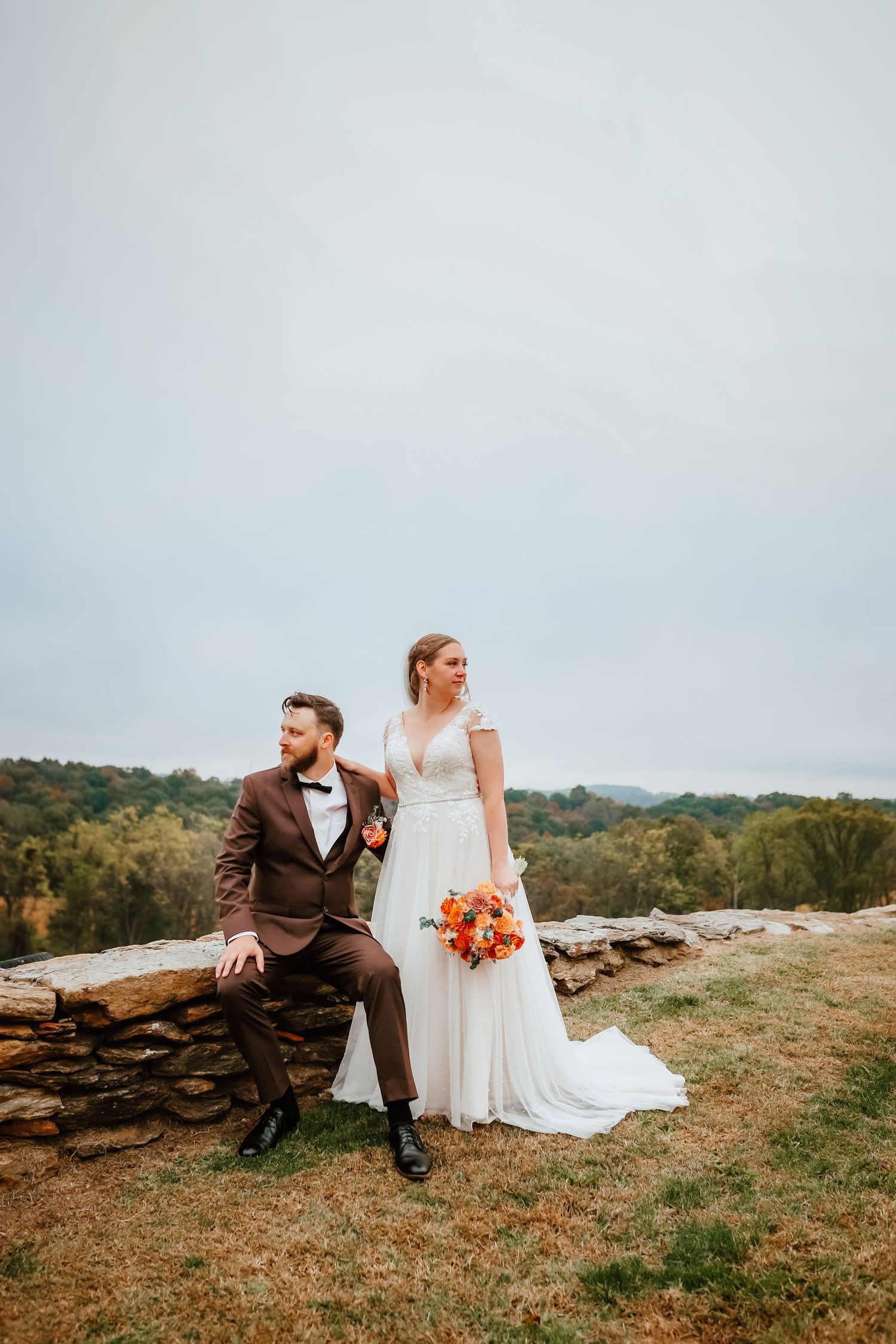 Bride and groom posing on stone wall with colorful autumn bouquet and mountain landscape backdrop.