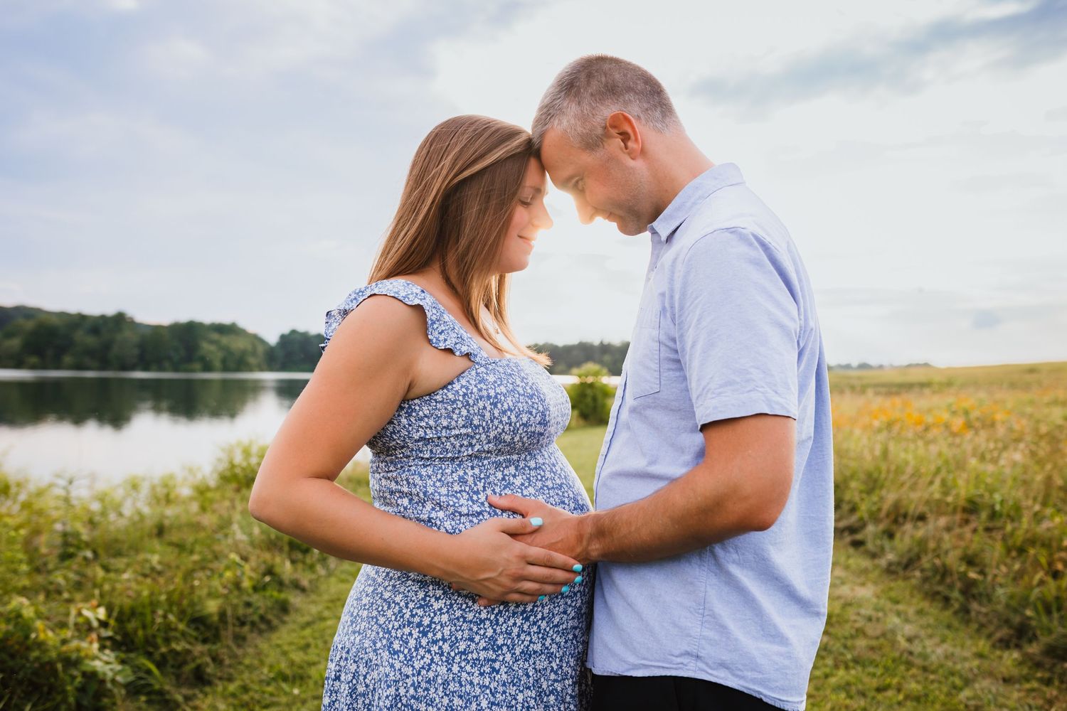 Pregnant couple standing by lake, facing each other with hands on baby bump under cloudy sky.