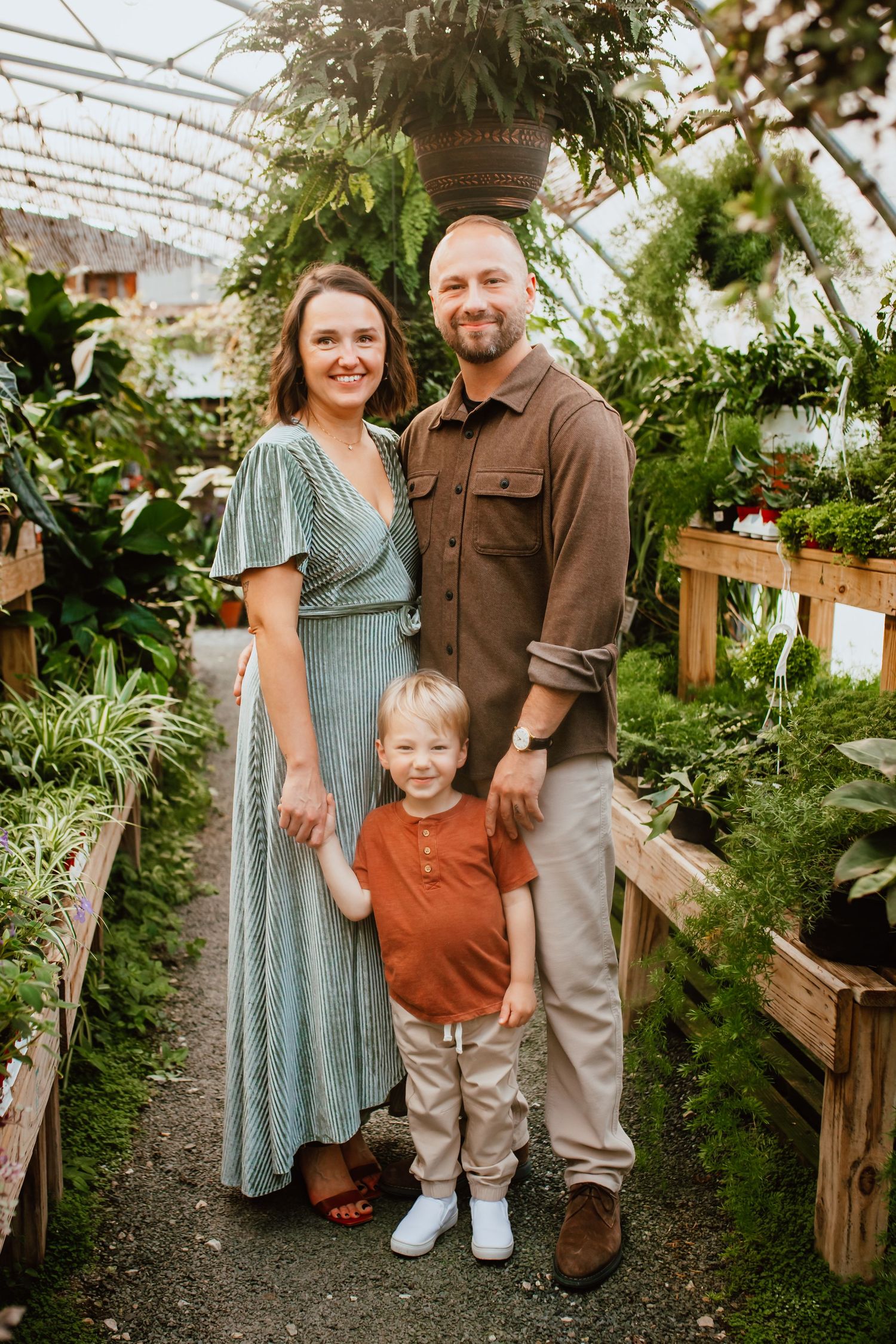 A smiling family of three poses in a lush greenhouse, with parents flanking their young son among vibrant green plants.