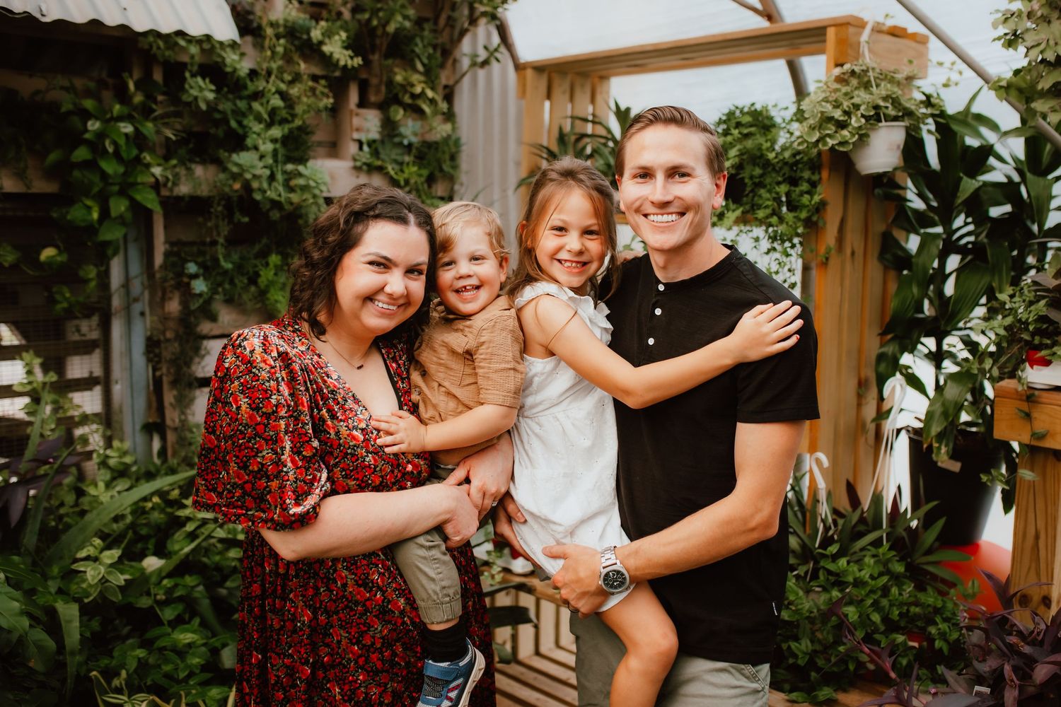 Happy family of four smiling together outdoors surrounded by lush green plants and wooden pergola.