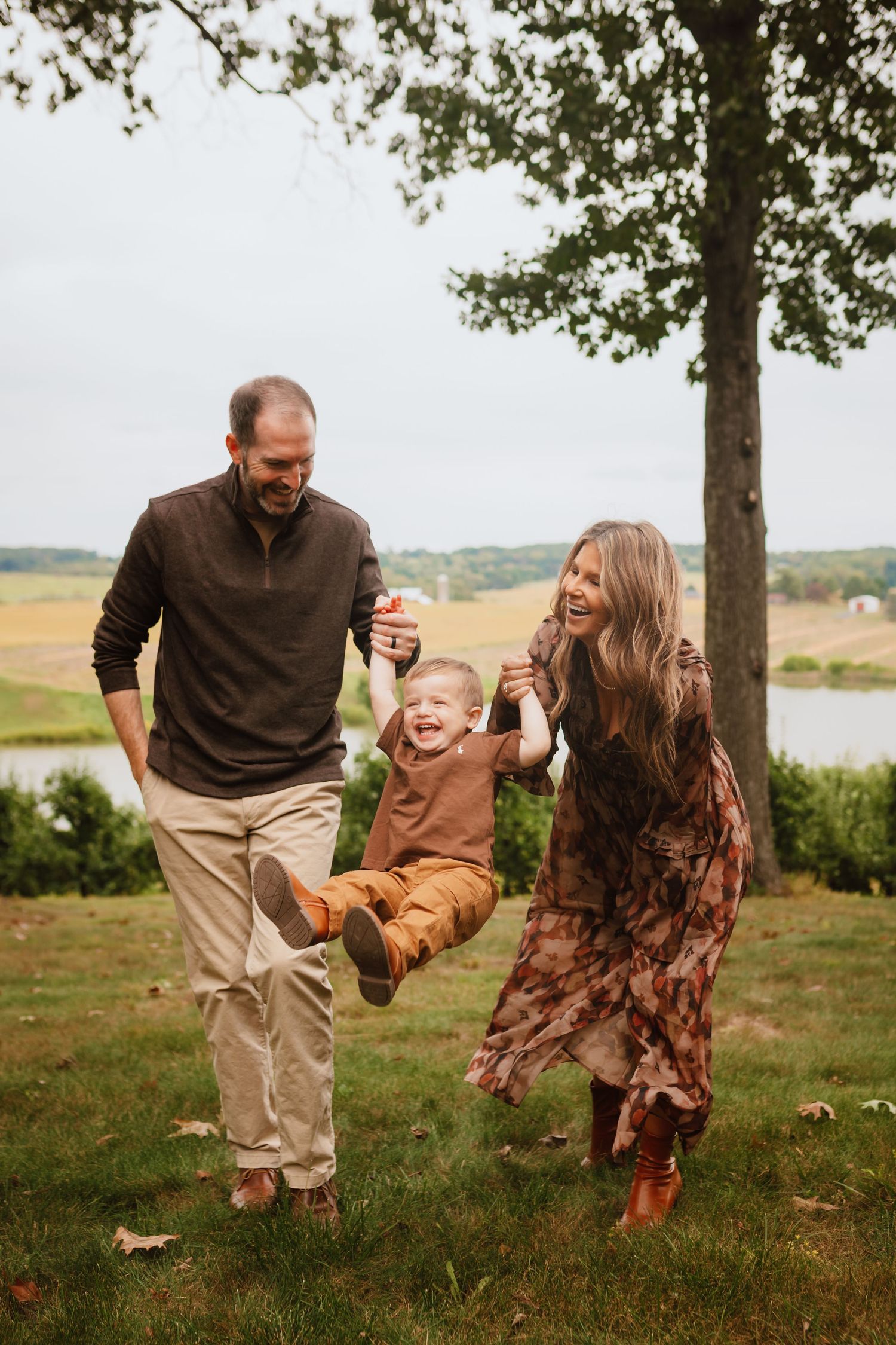 Smiling parents swing their laughing toddler by the hands on a grassy lawn beside a tree, with a calm lake and fields behind.