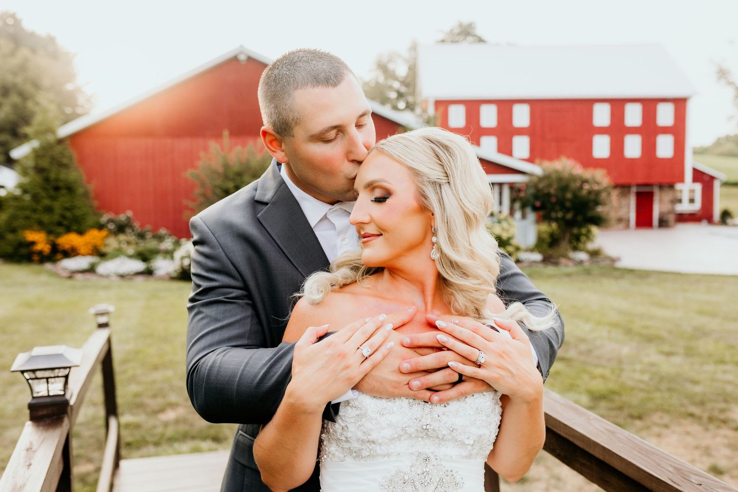 Groom kisses bride on the cheek as they pose in front of a classic red barn on their wedding day.
