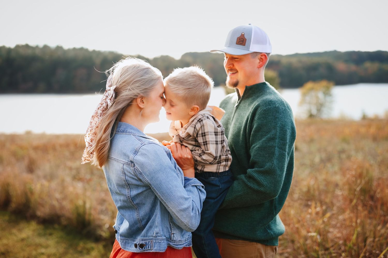 Young couple holding their baby boy outdoors near a serene autumn lakeside.