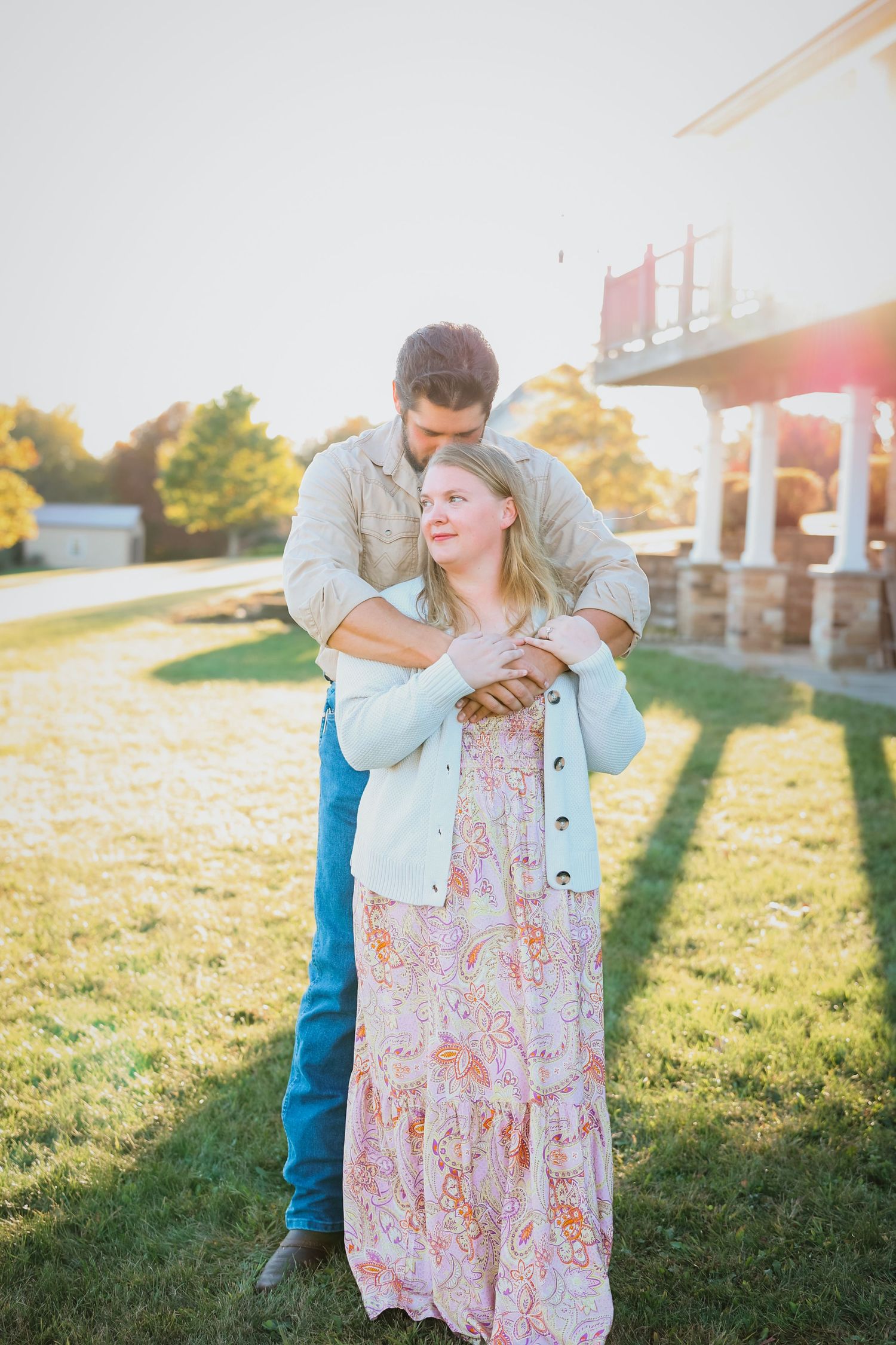 Couple embraces in golden sunset light on a green lawn beside a white building.