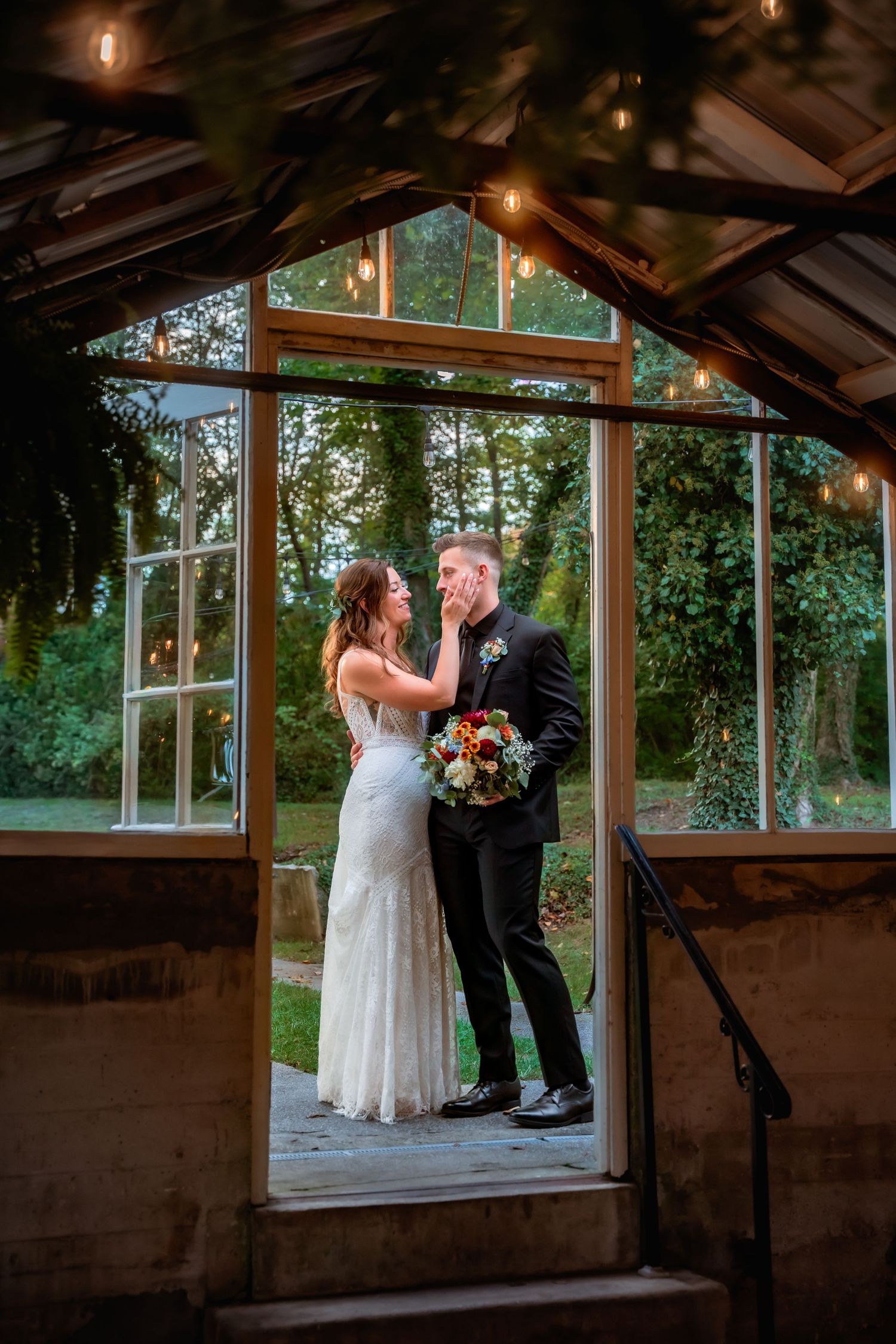Bride and groom share a romantic moment inside a glass greenhouse adorned with string lights.